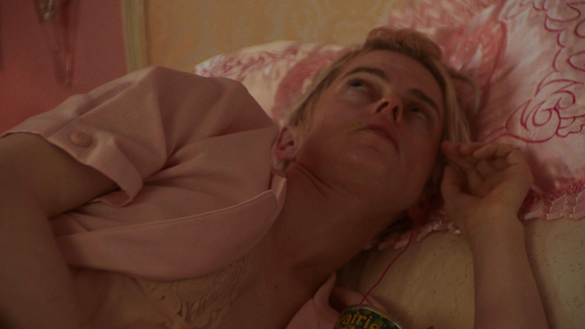 A woman lying in a pink-themed bedroom eats canned peas alone while another individual engages in a conversation via a smartphone nearby. The atmosphere conveys a sense of quiet isolation and personal routine.