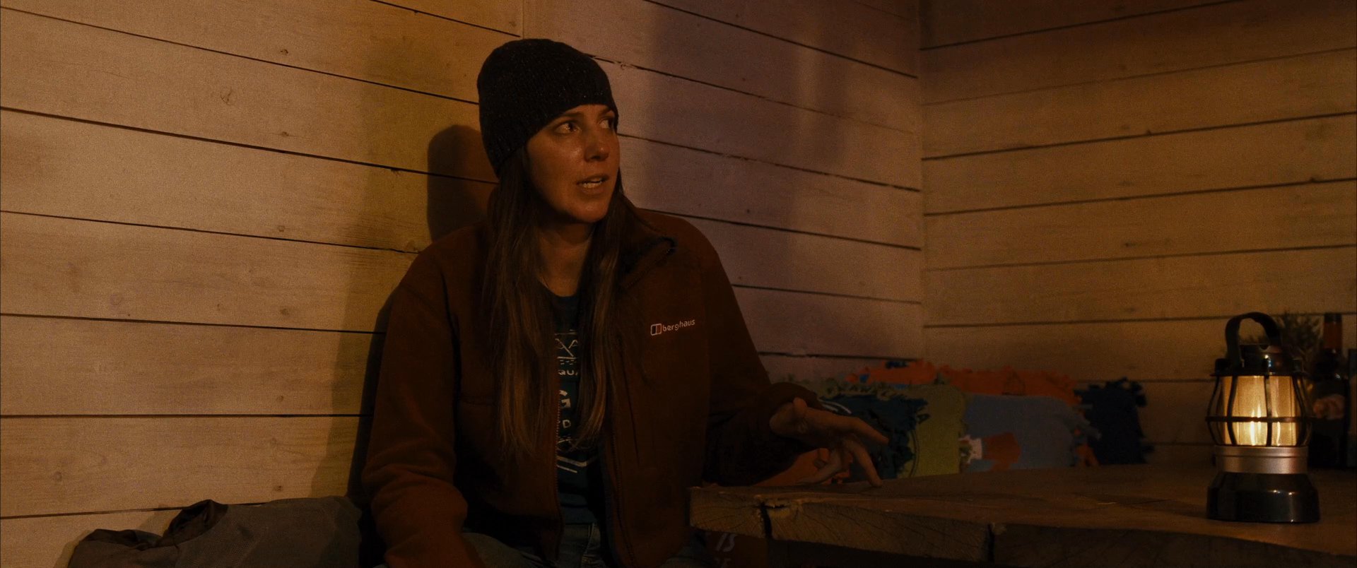 Two individuals engage in a focused conversation while seated inside a dimly lit cabin illuminated by a lantern. The scene emphasizes their interaction as they exchange dialogue in a quiet, rustic environment.