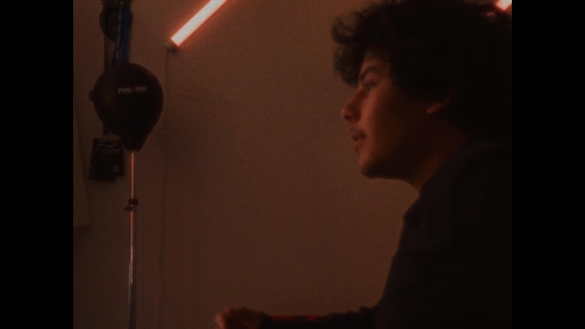 A young man delivers a direct-to-camera monologue in a dimly lit bedroom setting, punctuated by close-up shots and expressive shifts in demeanor. The warm, ambient lighting emphasizes an introspective and engaging atmosphere.