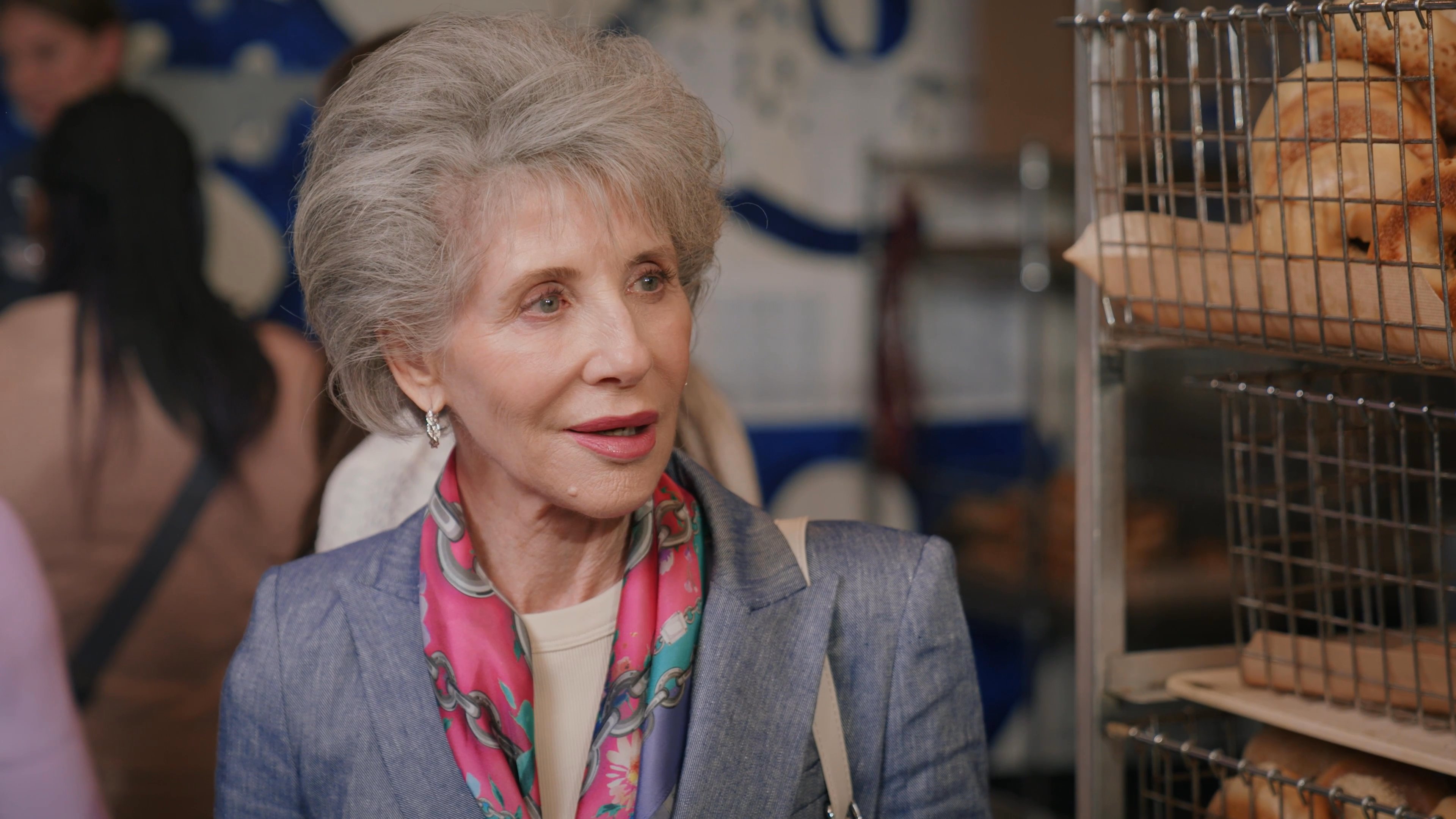 An elegant older woman engages in a polite conversation within a bakery setting, surrounded by shelves stocked with bread and pastries.
