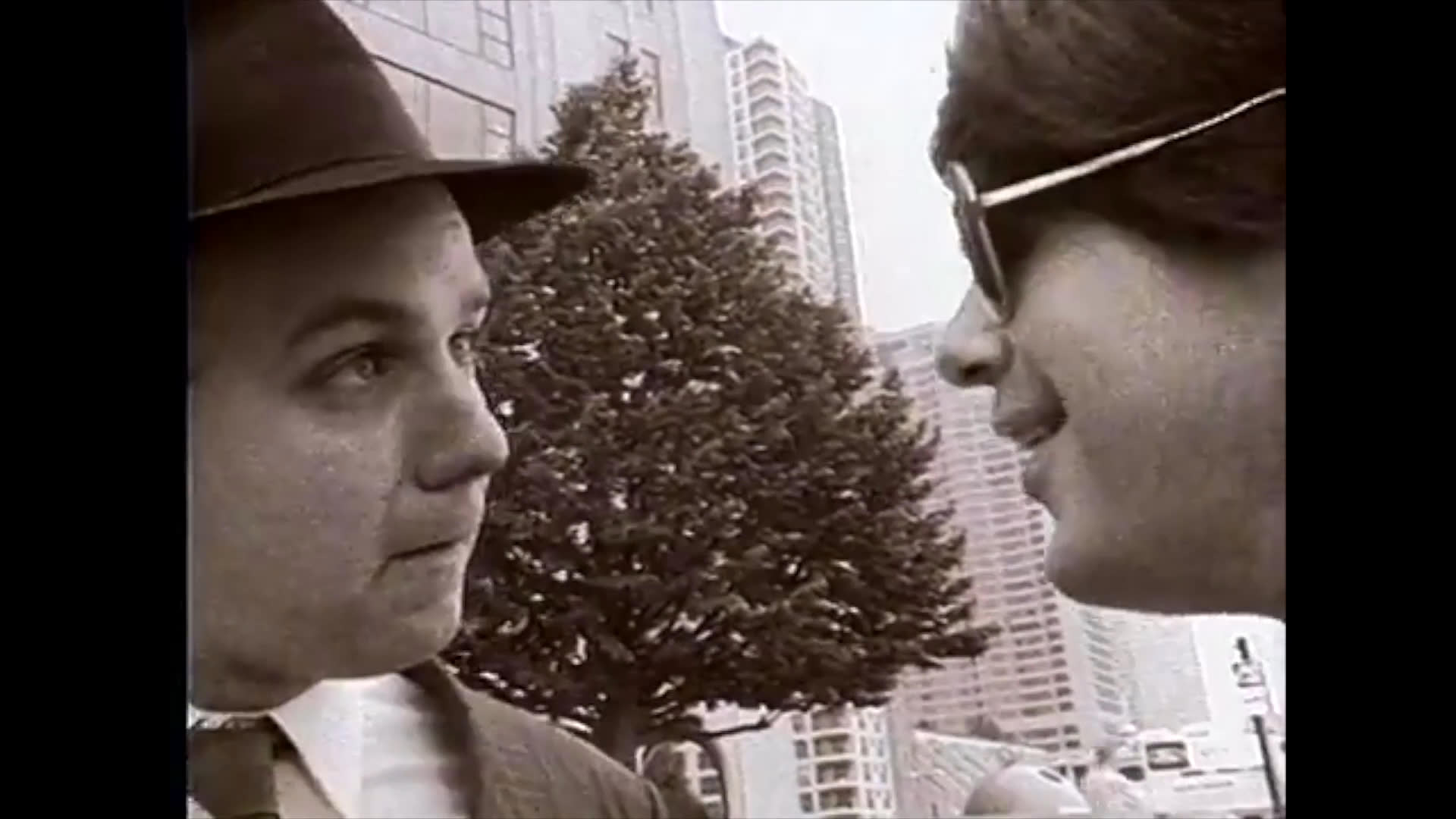 Two men engage in a conversation while standing on an outdoor pier against an urban backdrop, later transitioning to a scene within a retail environment. The exchange reflects a professional yet informal narrative style captured in a classic monochromatic aesthetic.