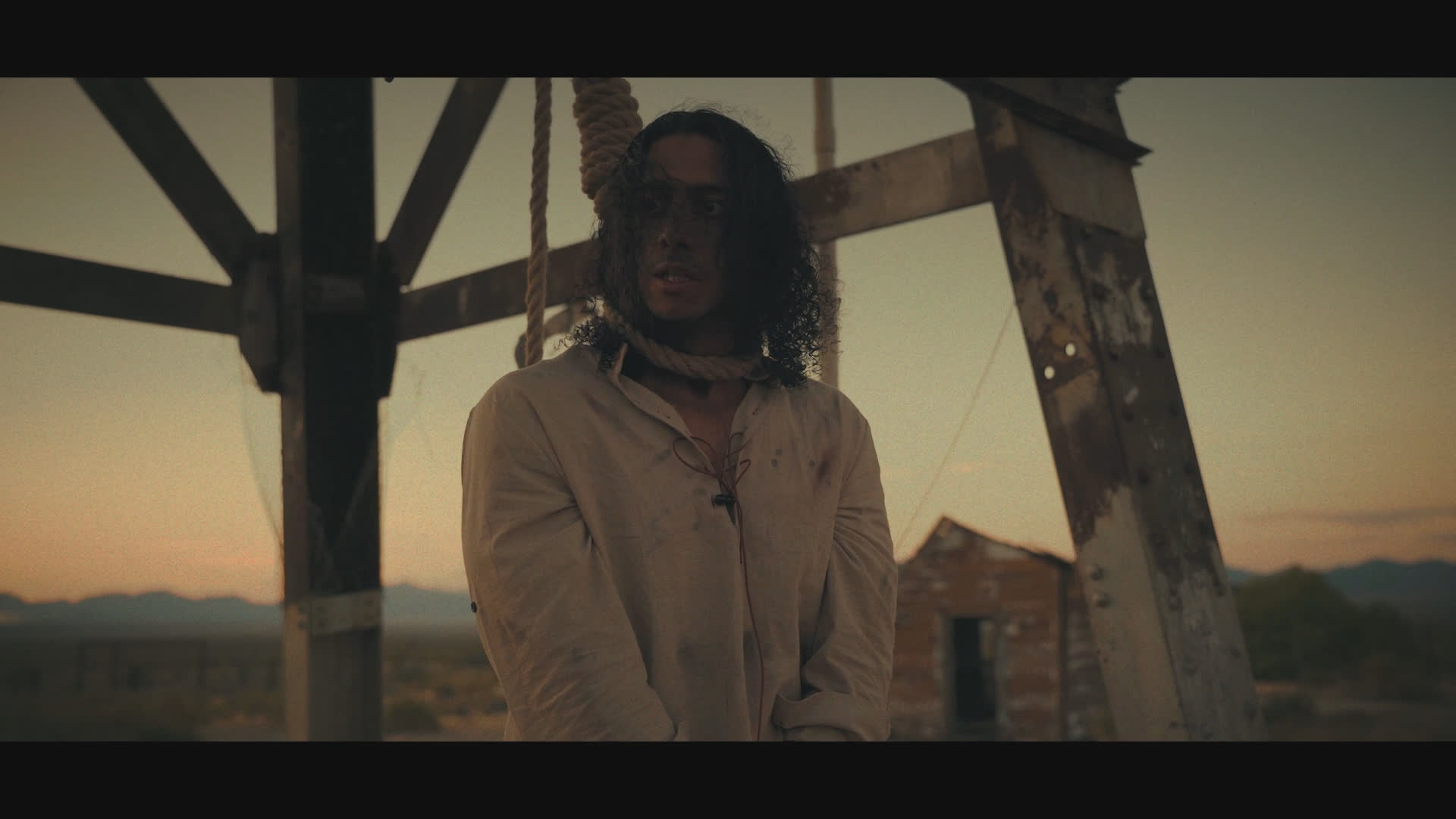 At sunset in a weathered frontier settlement, a hat-wearing figure gestures before the scene shifts to a long-haired man standing beneath a wooden gallows-like frame. Their tense exchange includes a remark about trying to kill himself.