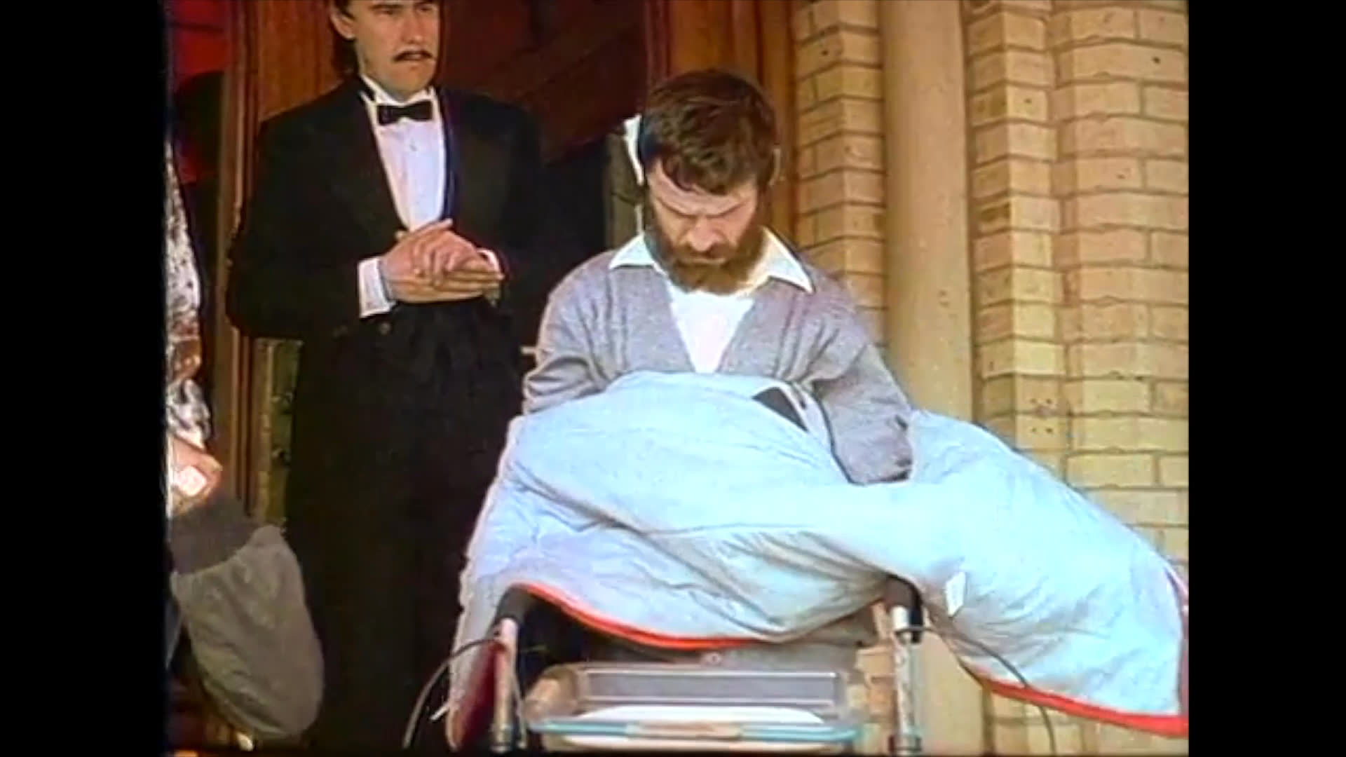 A man meticulously puts on a multi-layered jacket while standing in a formal setting, observed by an attendant.