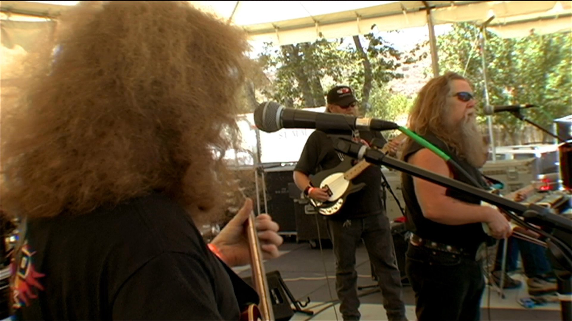 A long-haired, bearded guitarist and vocalist performs live on an outdoor stage at a musical event. He sings into a microphone while accompanied by bandmates in a sunny, tented setting.
