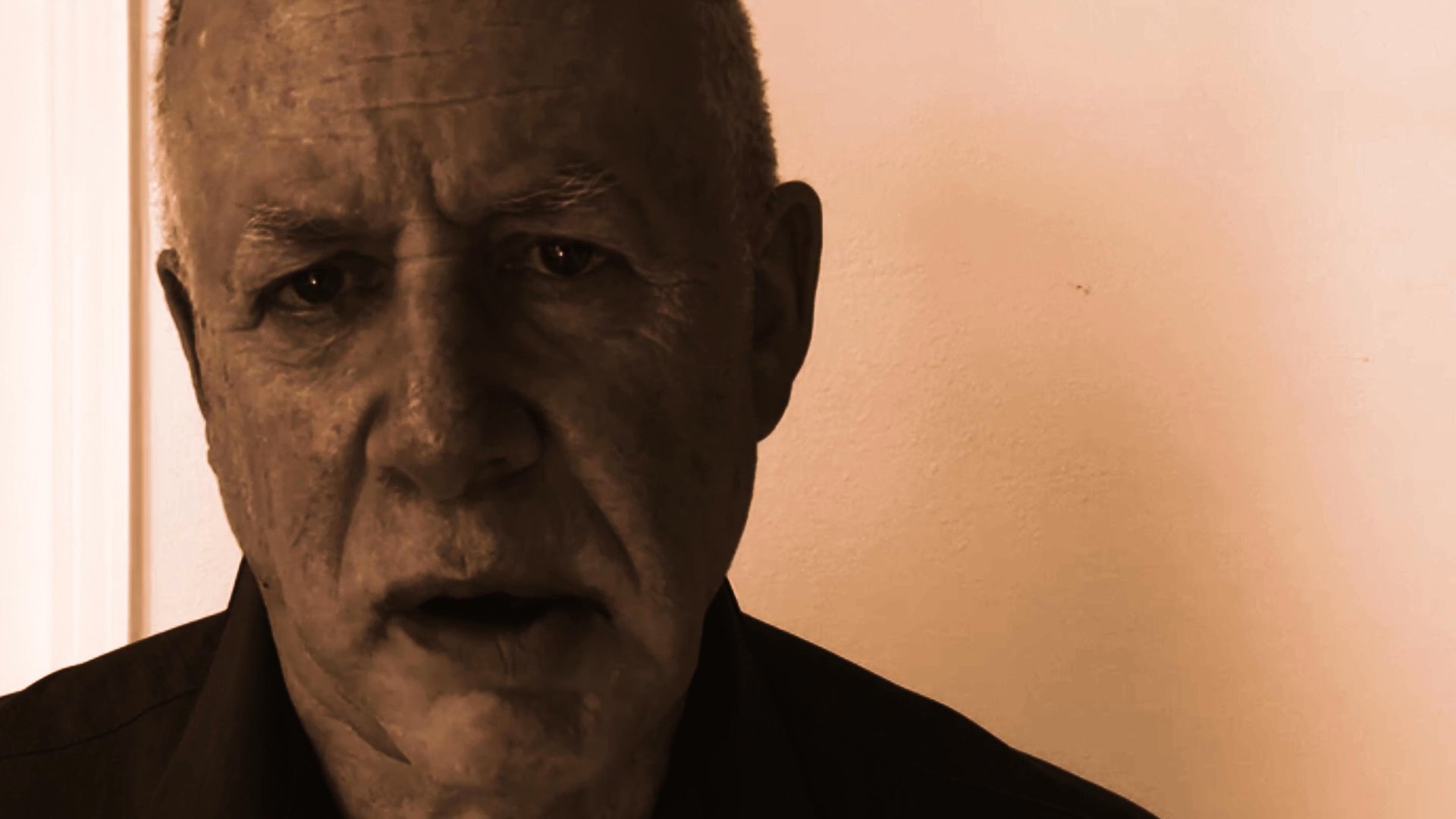 An older man delivers a direct address to the camera in a sepia-toned, close-up shot. He maintains steady eye contact while speaking throughout the sequence.