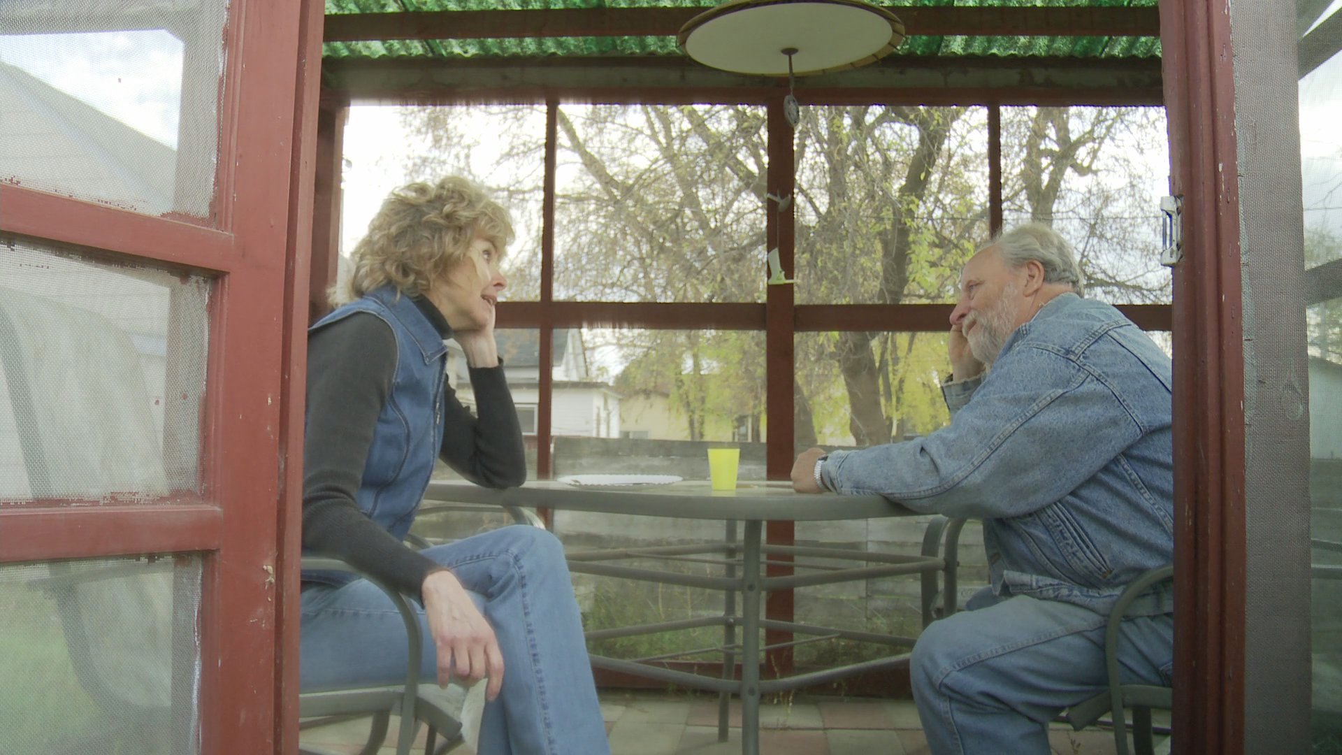 A mature couple engages in an intimate conversation while seated inside a sunroom, transitioning from a formal dining setting to a quiet outdoor enclosure.