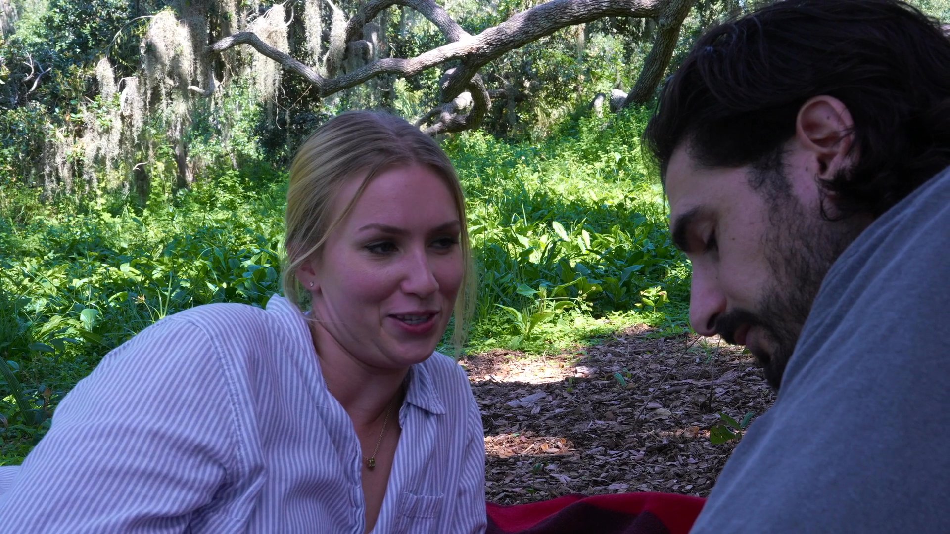 A man and a woman share an intimate conversation while sitting on a blanket beneath a large, sprawling tree in a park. The scene highlights their affectionate engagement in a serene, natural environment.
