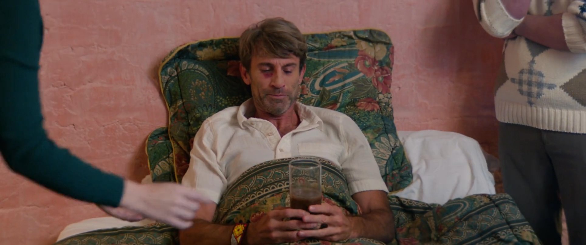 A group of caregivers provides support and delivers a drink to a man resting in bed. The scene captures a moment of attentive care within a residential bedroom setting.