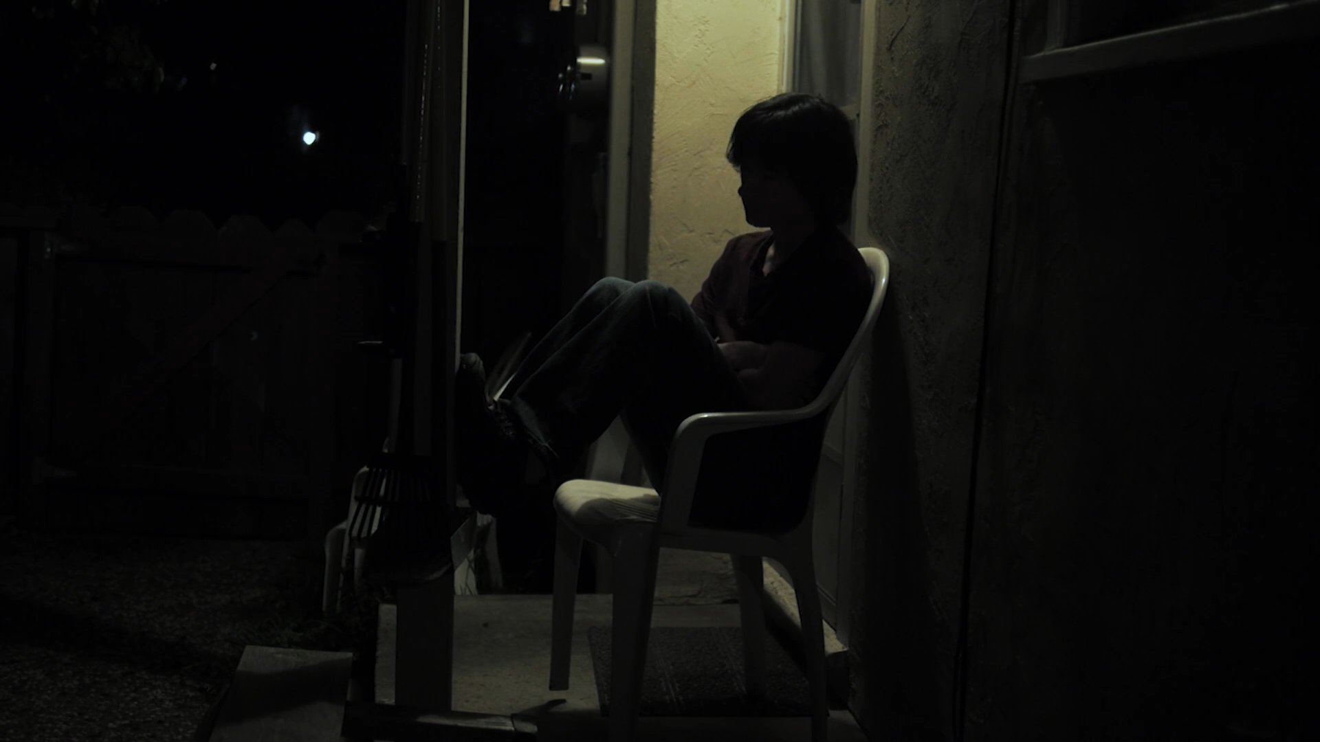 A woman sits alone on a chair outside against a wall at night, appearing lost in thought or quiet reflection.