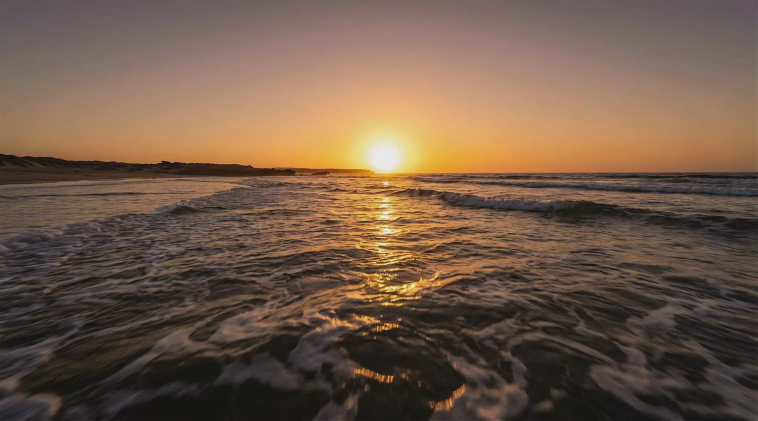 A wide-angle video frame showcasing a sunset over the ocean, generated by Kling 2.5 Turbo at 1928x1072 resolution. Gentle waves with white foam ripple in the foreground, reflecting the bright golden sun low on the horizon under a clear, warm gradient sky.