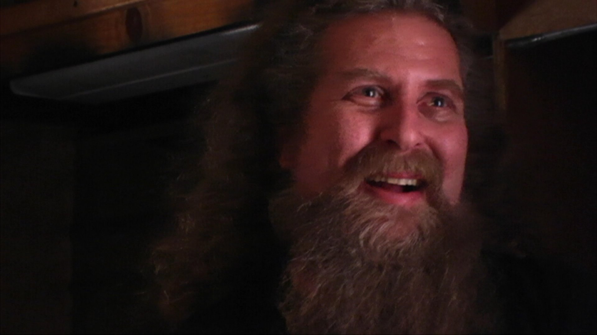 A man with a long beard and long hair shares personal experiences and anecdotes during a candid, dimly lit interview session.
