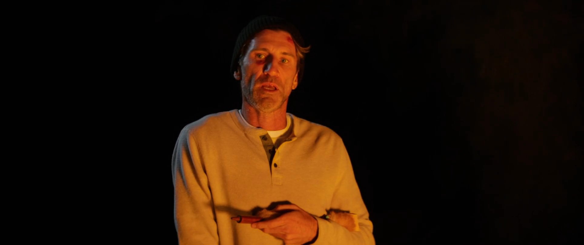 A man wearing a beanie and henley shirt speaks to a small group gathered around a fire in the darkness. The warm glow of the flames illuminates his face as he narrates, creating an intimate evening atmosphere.
