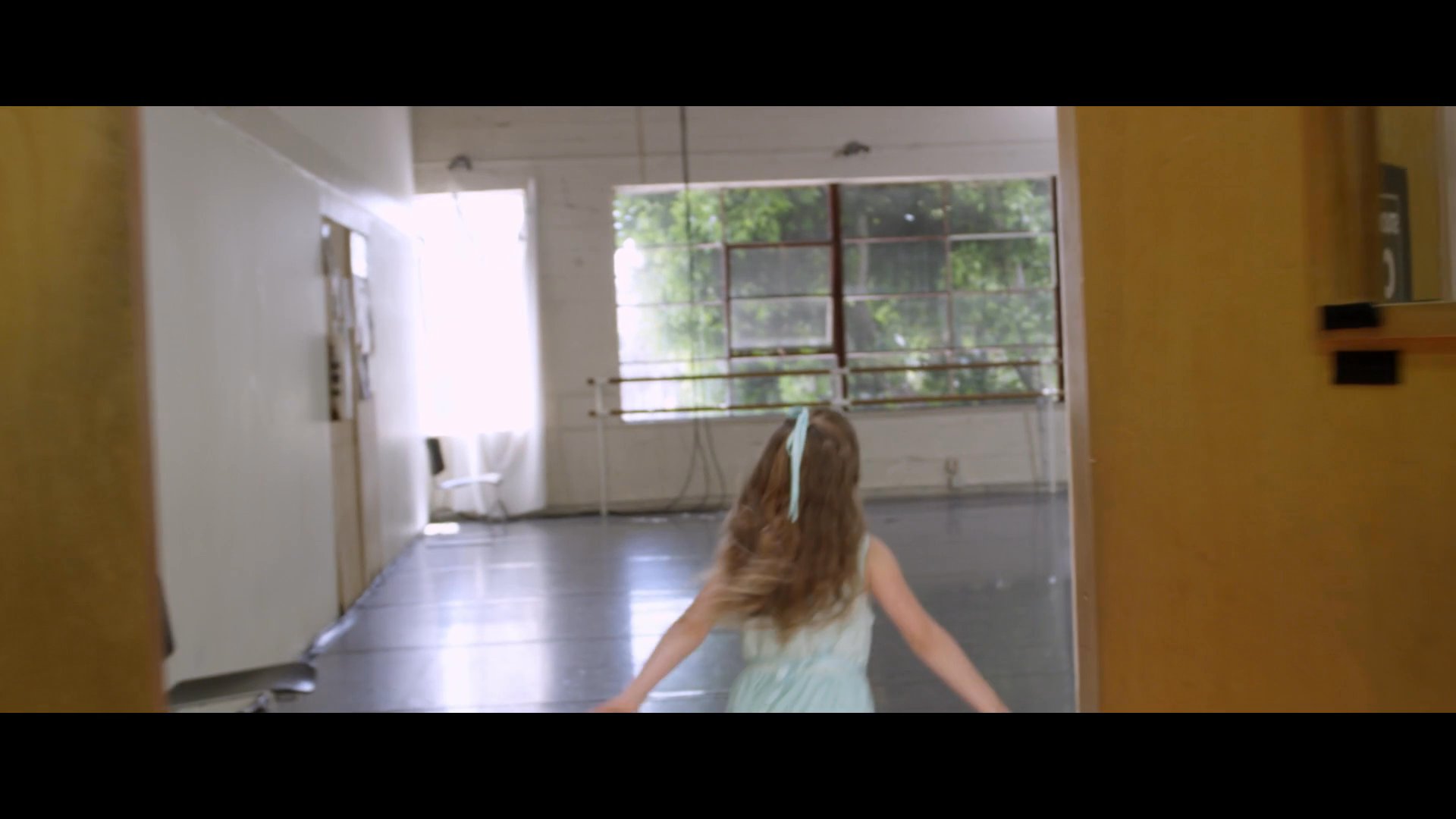 A young student enters a professional studio to join a group of dancers in a classical ballet training session. The class practices choreographed movements under the guidance of an instructor.