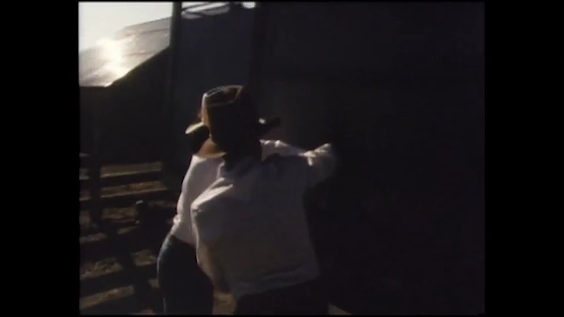 Two people stand beside a large structure on a rural property, gesture toward it, and walk away as a vehicle heads down the country road. The exchange feels reflective, accompanied by a voice recalling a life well lived.