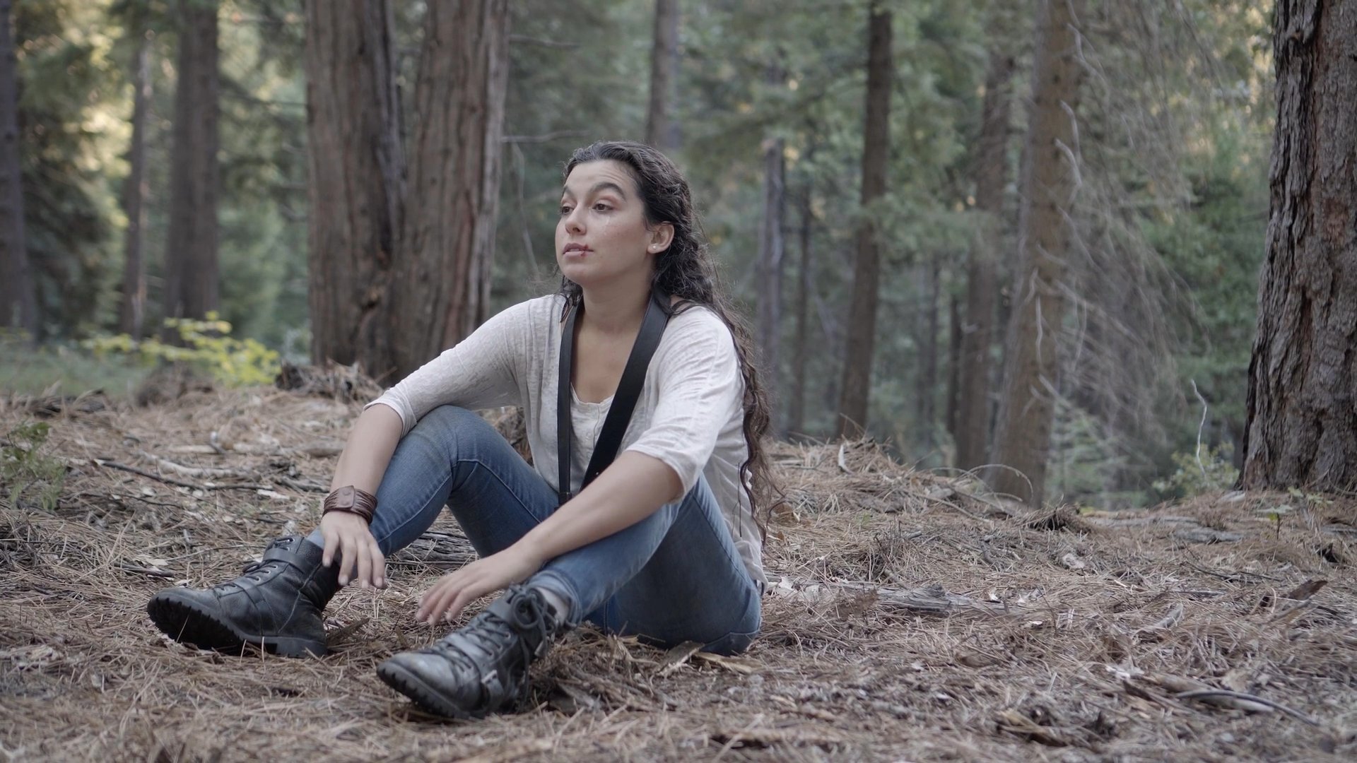 A woman sits cautiously in a dense forest as she notices a mysterious creature with glowing eyes observing her from a tree stump.