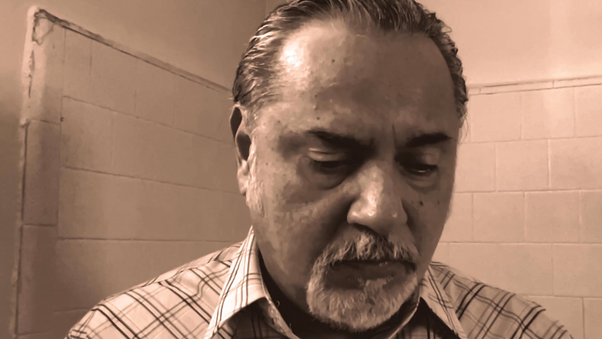 A man with a grey beard speaks directly to the camera in a sepia-toned, indoor setting. He delivers a focused message while maintaining steady eye contact.