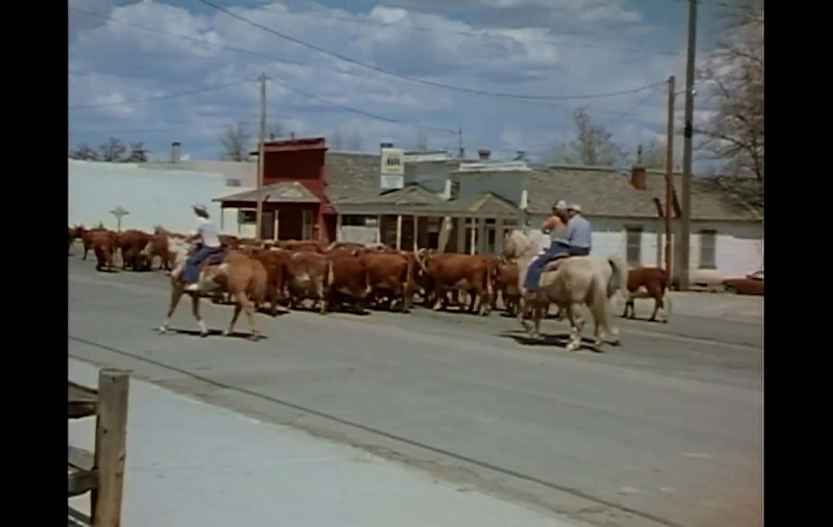 A herd of cattle moves across a small-town main street as riders guide them past storefronts and houses. Snow-capped mountains rise behind the town as the cattle spread across the road.