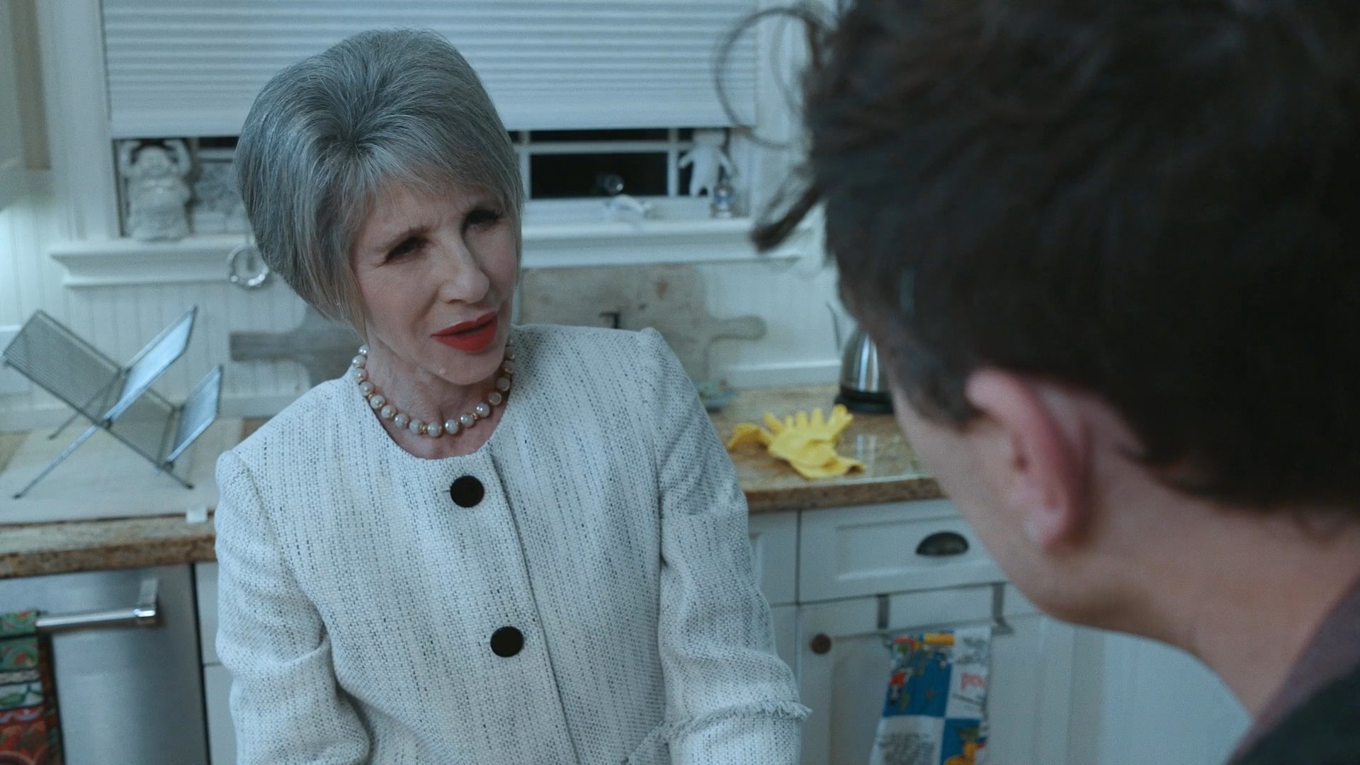 A man and an older woman engage in a serious, intimate conversation while sitting together in a home kitchen. The exchange captures a moment of emotional tension or significant discussion between family members.