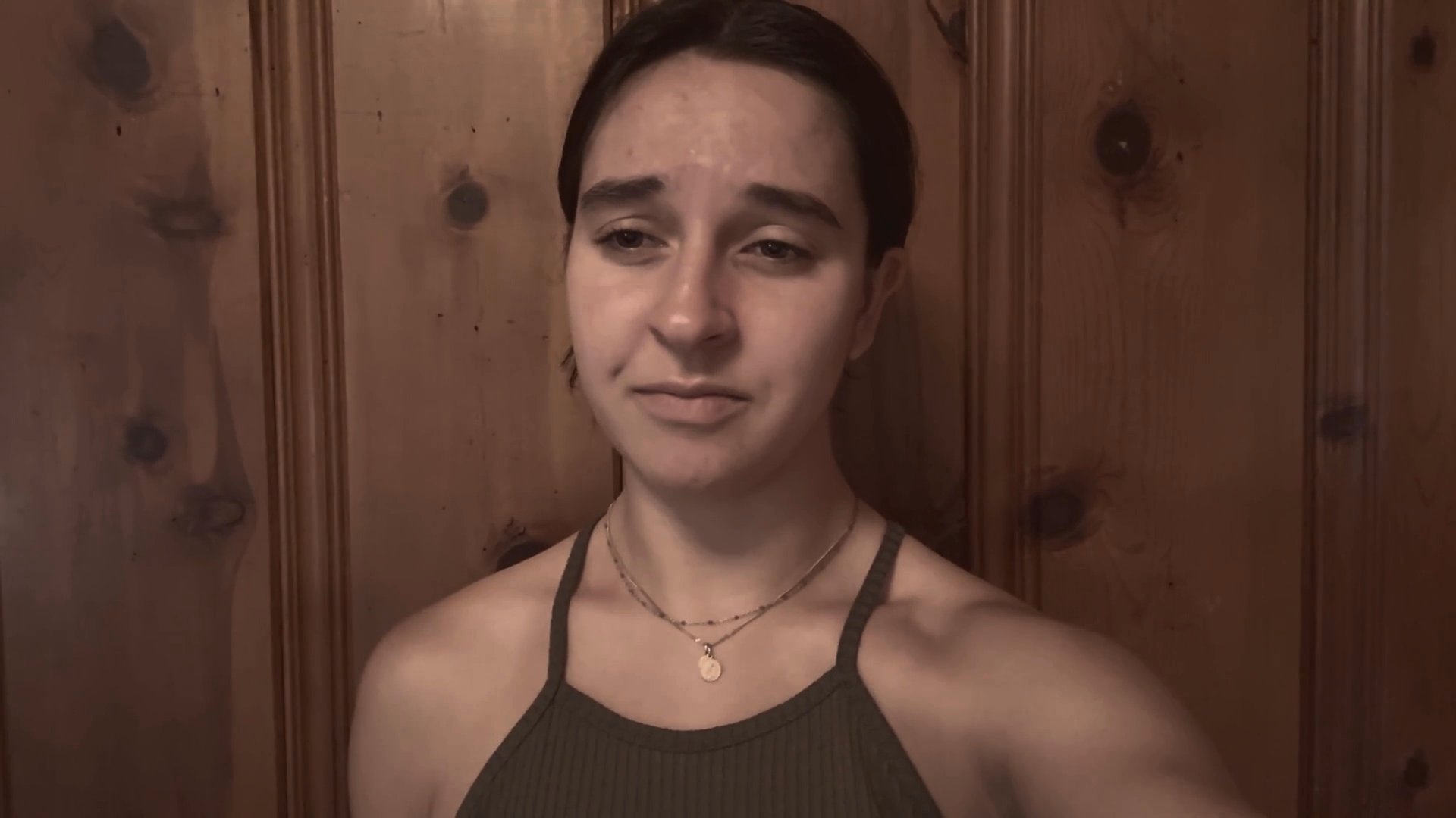 A woman stands before a wooden backdrop and speaks directly to the camera with varied facial expressions and emotive delivery. Her performance shifts between thoughtful reflection and direct address.