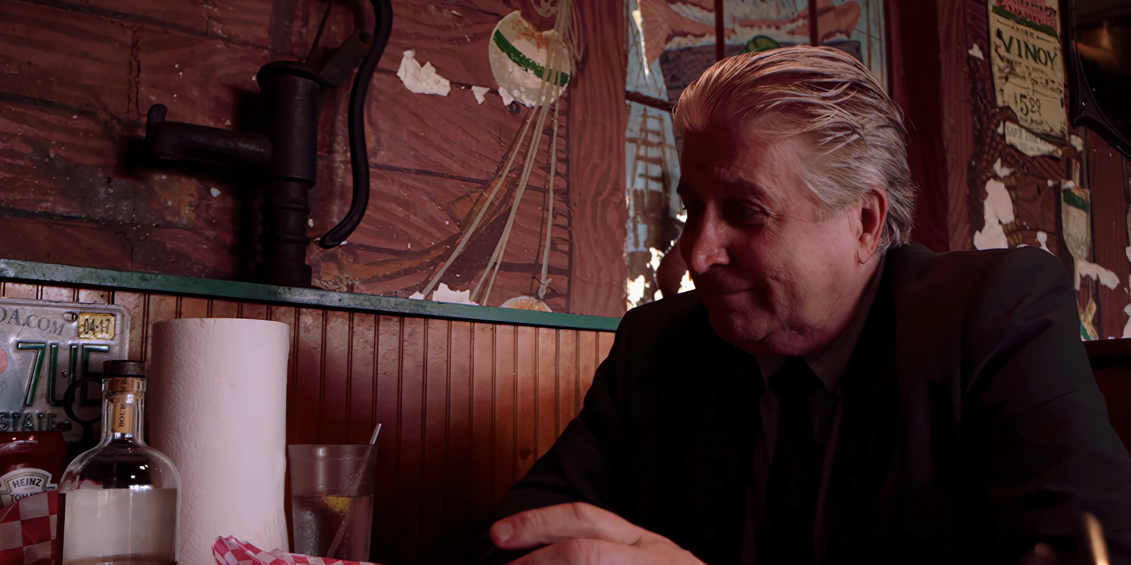 Two men engage in a serious discussion while seated in a wood-paneled diner booth. The scene emphasizes their interaction as they talk across the table in an intimate setting.