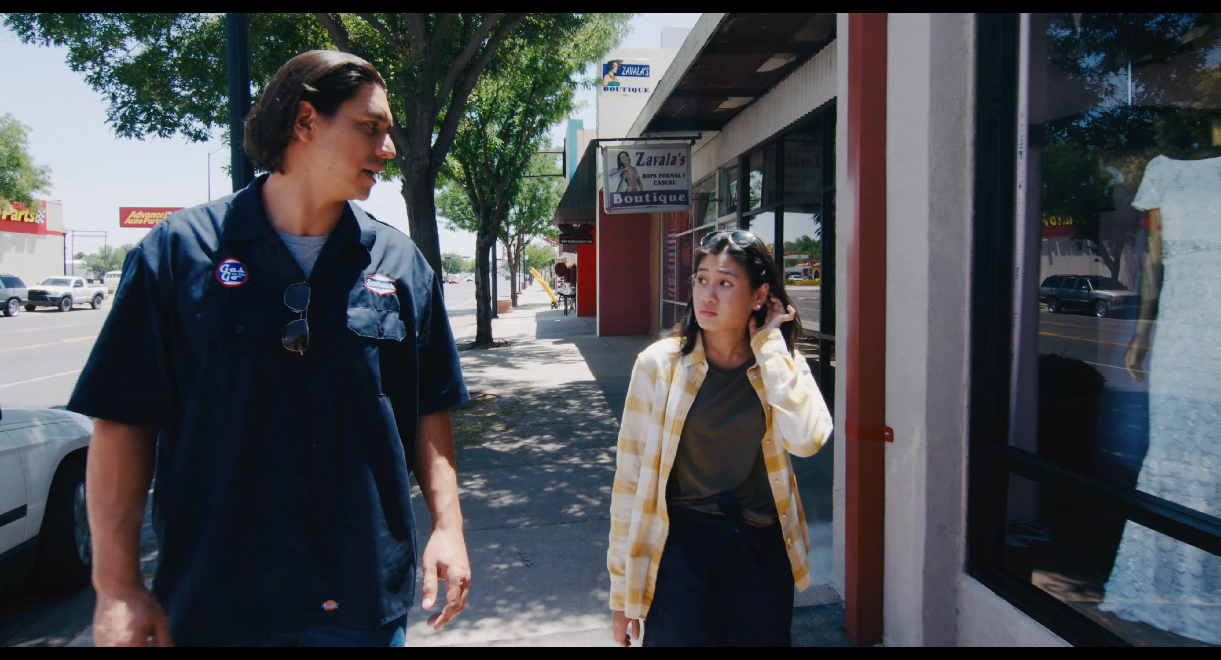 A man and woman engage in conversation while walking along a sidewalk past a local storefront.