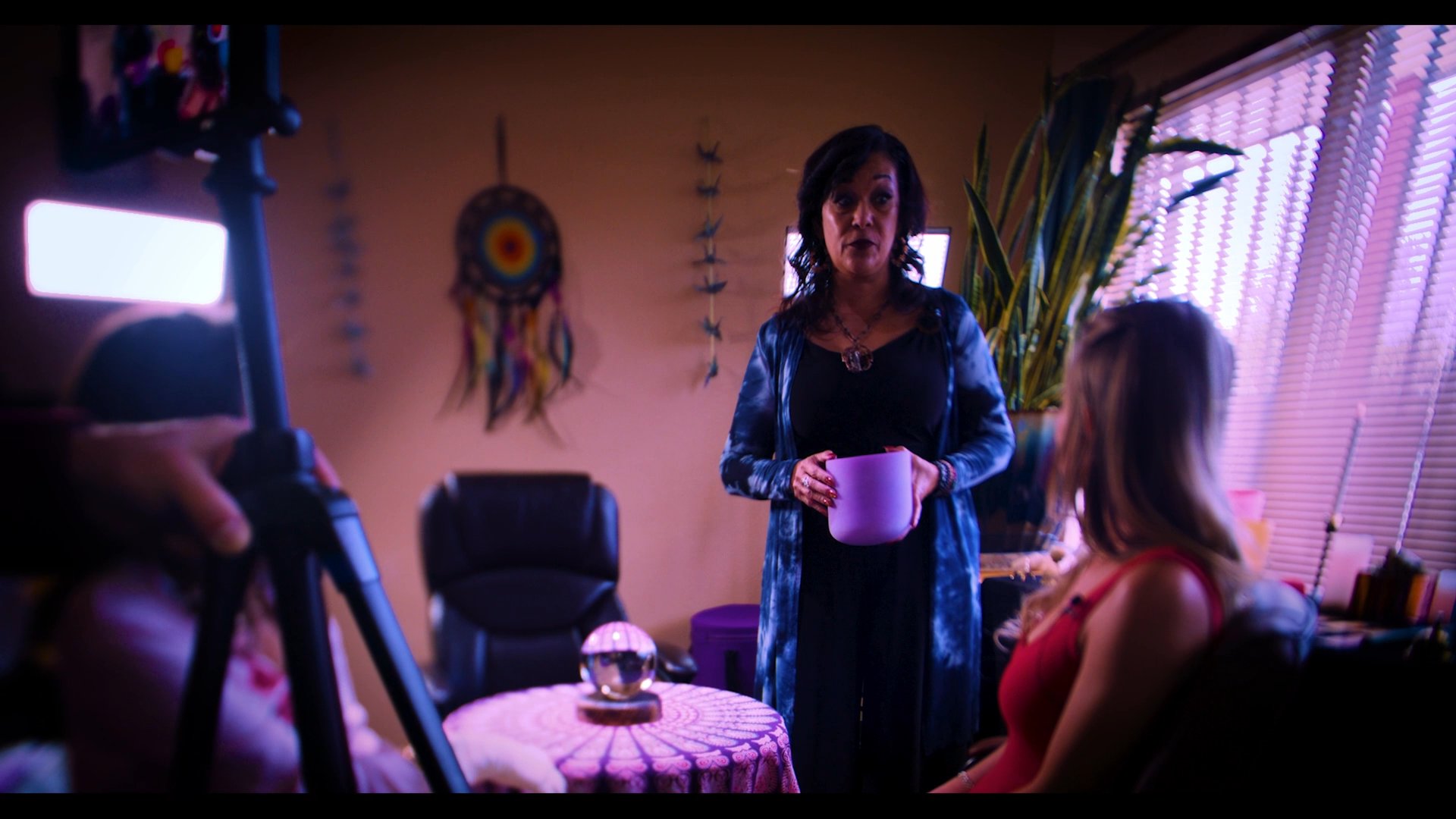 A woman receives a spiritual reading from a practitioner holding a purple container in a dimly lit, atmospheric room.