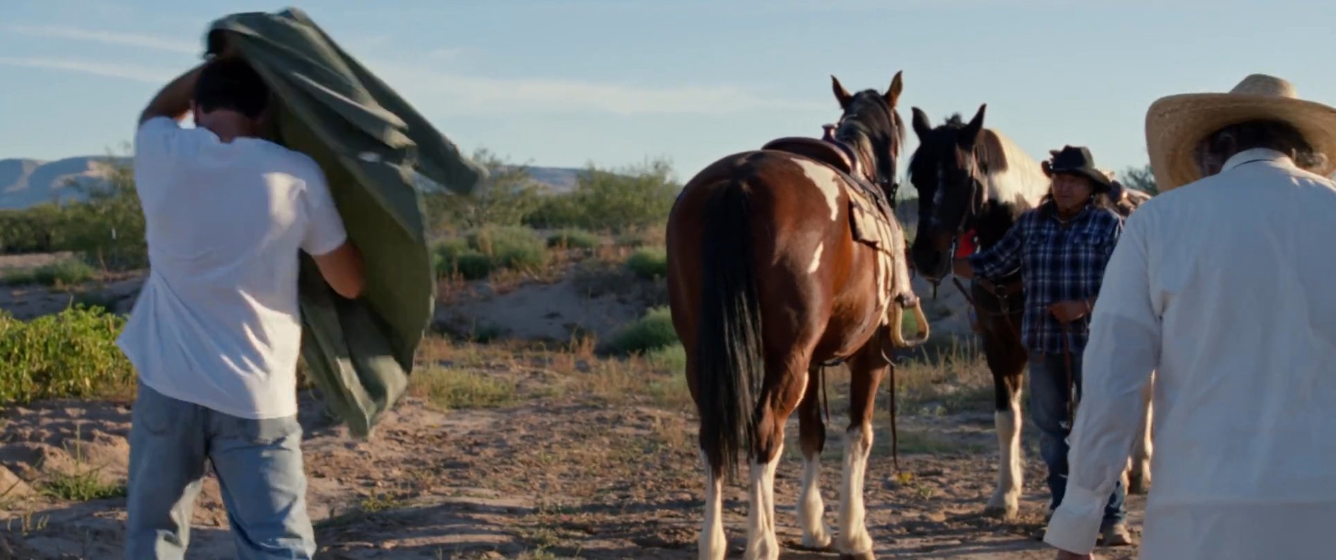 An experienced cowboy provides guidance and assistance to a novice rider as they prepare to mount a horse in a desert landscape.