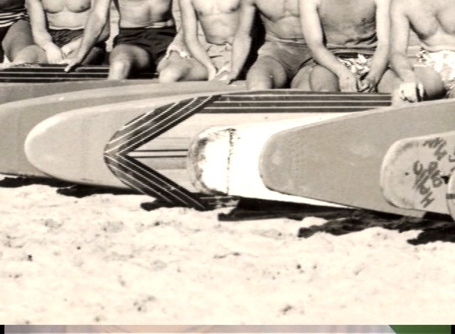 A group of surfers sits behind a row of wooden surfboards on a beach, posing for a group portrait.