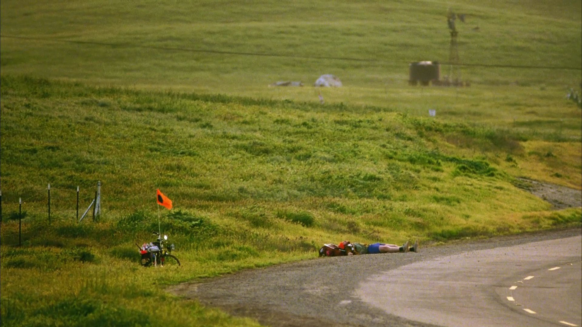 A backpacker signals for a ride along a rural road near a wind farm before resting on the grass. A passing car approaches, prompting the traveler to stand and resume signaling.
