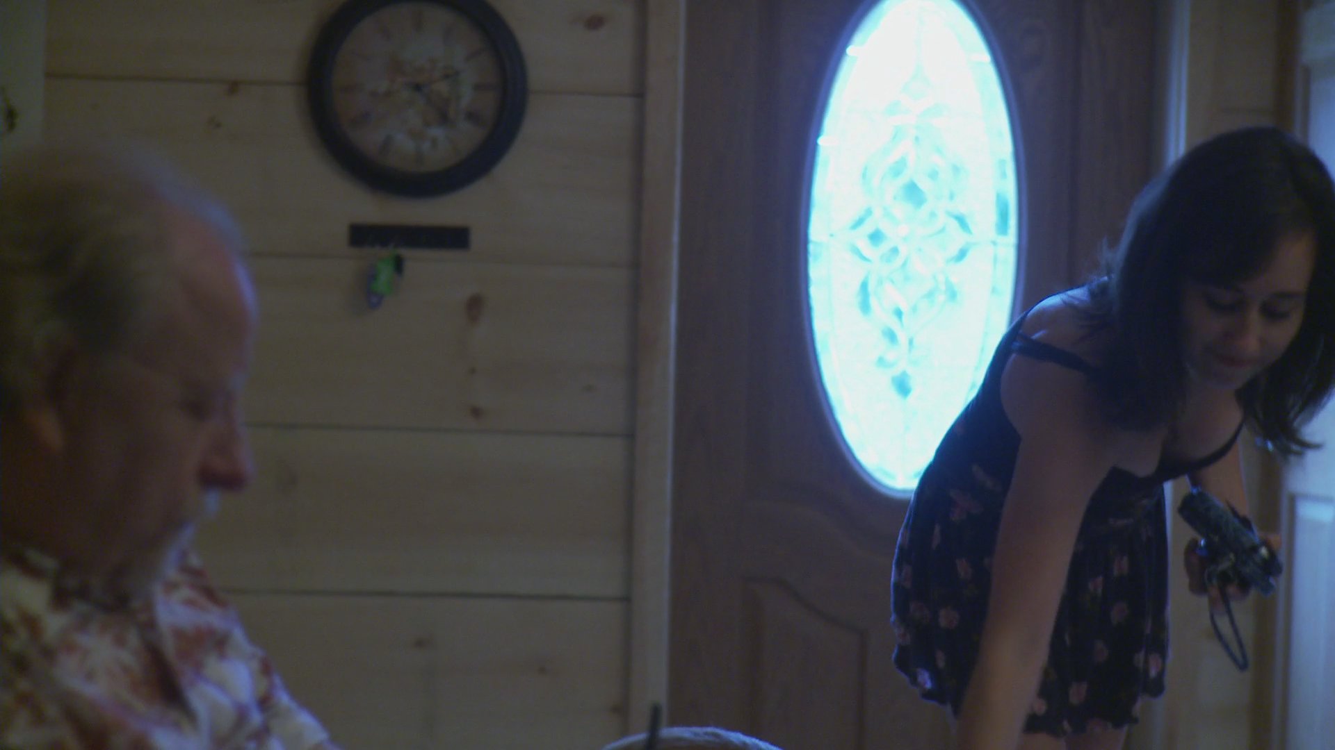 A woman enters a wood-paneled room and interacts with an older man, while another woman engages in activities in the kitchen area. The sequence captures a candid domestic scene within a residential environment.