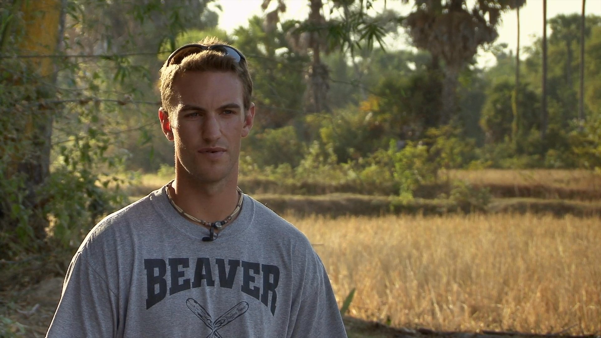 A man addresses the camera while standing in a rural, sunlit field. He maintains professional engagement during what appears to be a filmed interview segment.