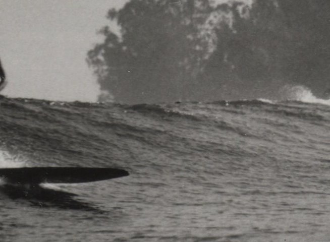 A surfer rides a longboard across a wave in a black and white coastal scene. The sequence highlights the fluid motion of the ride against a sunlit shoreline.