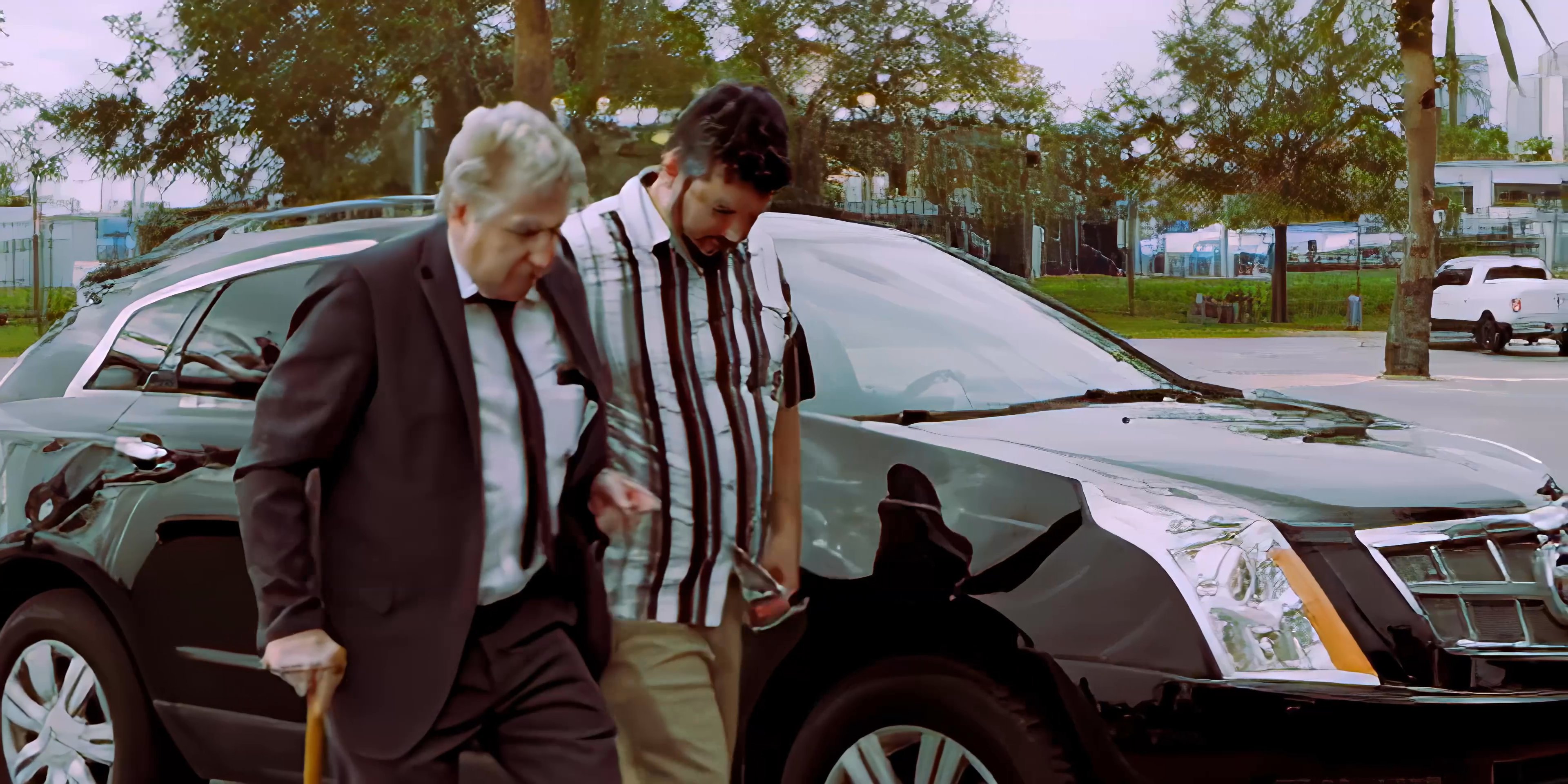 A younger man assists an elderly gentleman with a walking cane as they travel from their vehicle to a park bench for conversation.