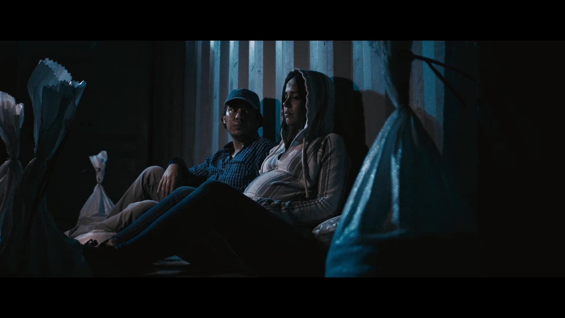 A man and woman sit in a dimly lit, confined space, sharing an intimate conversation while reflecting on their situation. The somber atmosphere emphasizes their proximity and shared focus during a tense moment.
