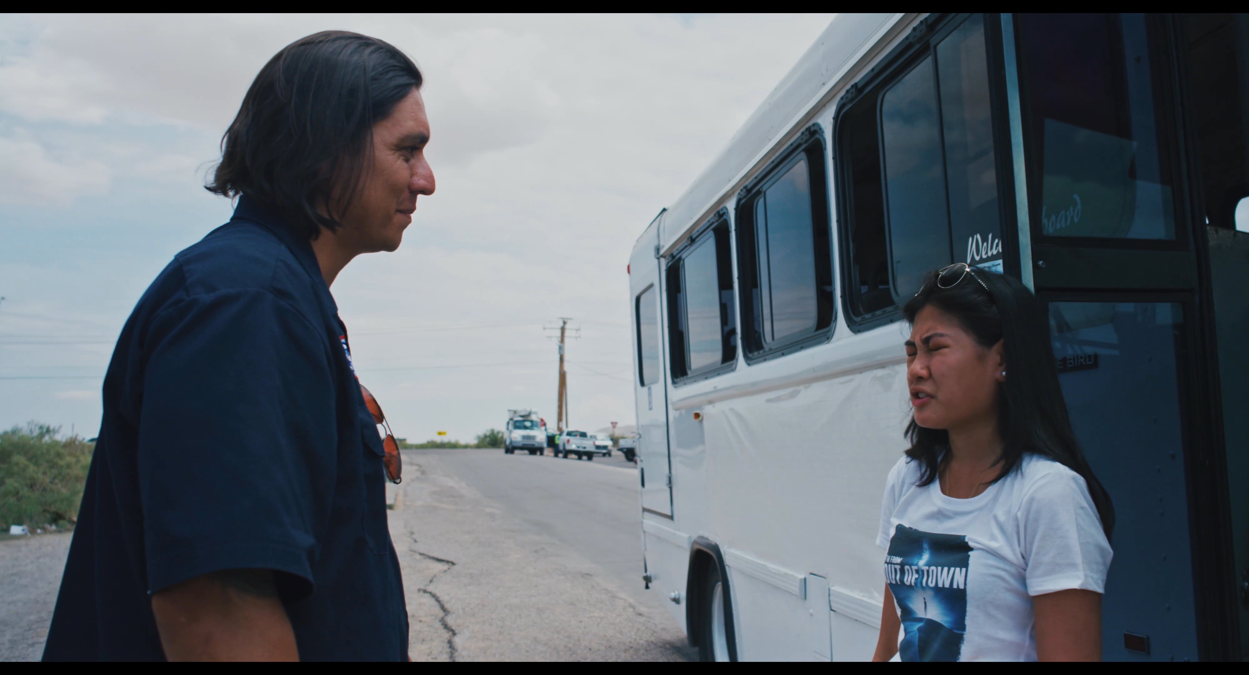 A man and a woman engage in a final conversation beside a parked bus before sharing an emotional embrace.