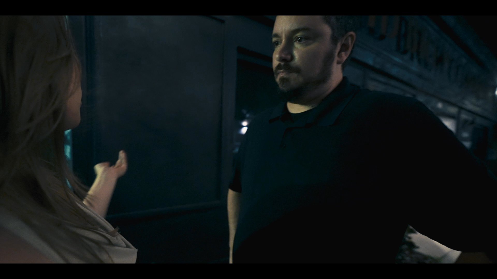A woman confronts a man outside a building at night, displaying visible frustration and physical tension during their heated interaction.