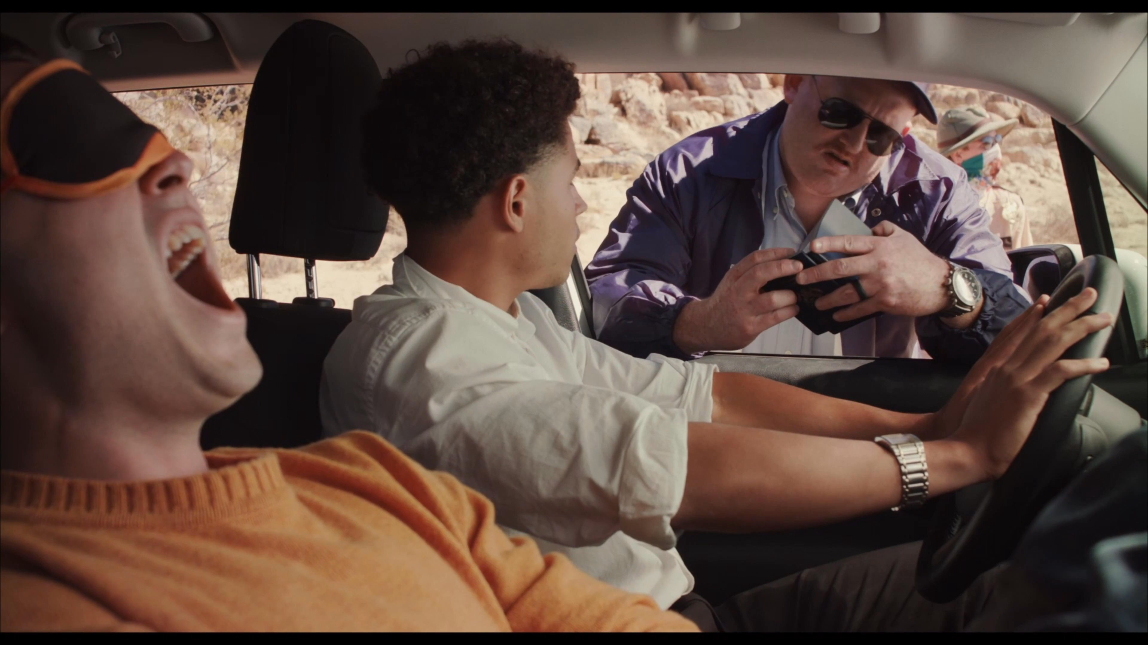 An officer in a desert setting questions two individuals inside a parked vehicle, resulting in a tense and expressive verbal confrontation.