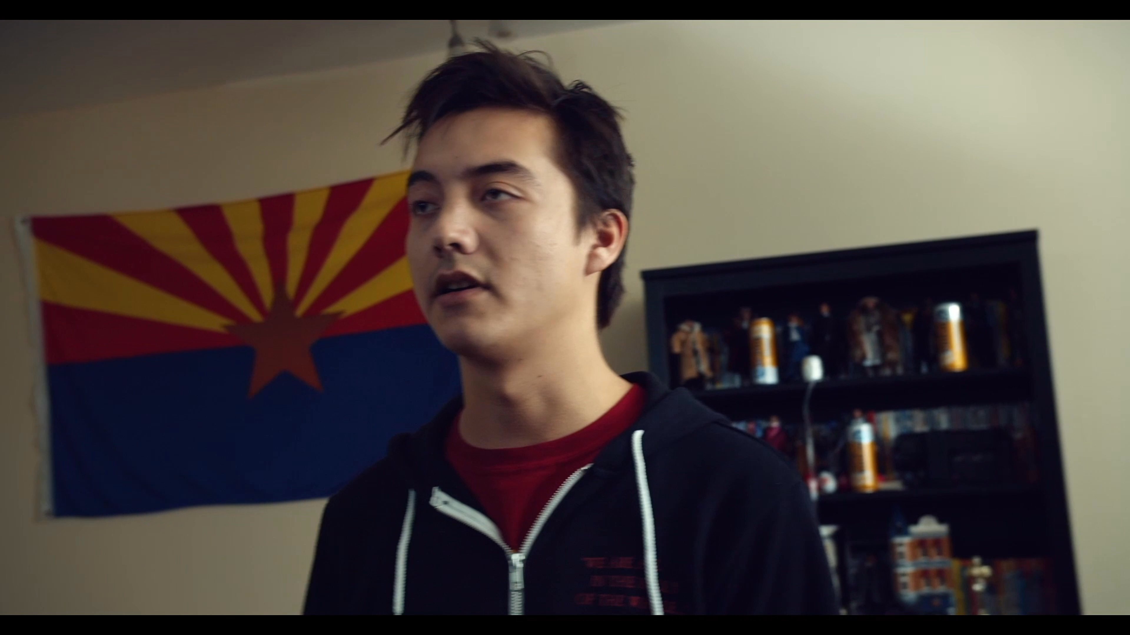 Two young men collaborate to pin photographs onto a corkboard, analyzing evidence in an informal apartment setting.