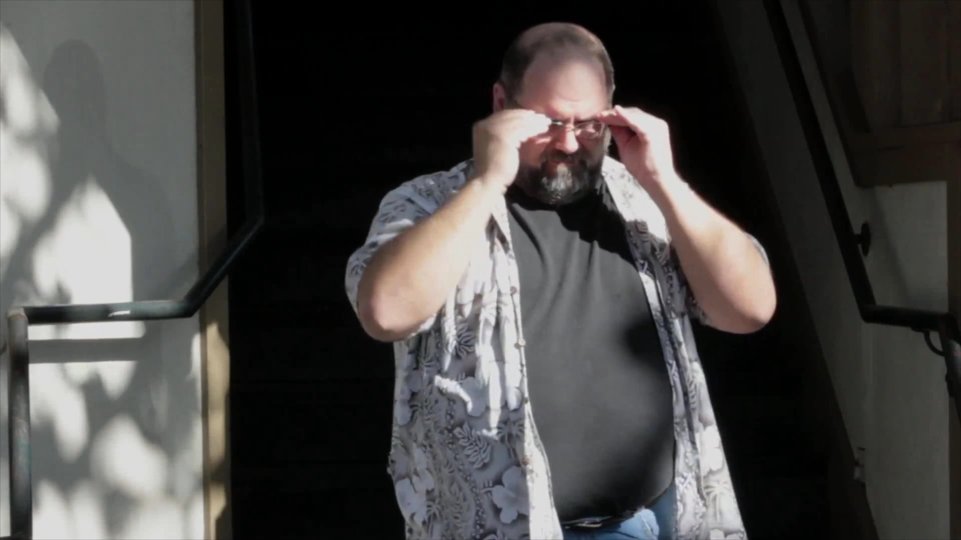 A man stands near an outdoor stairway, removing his glasses to clean them before placing them back on his face.