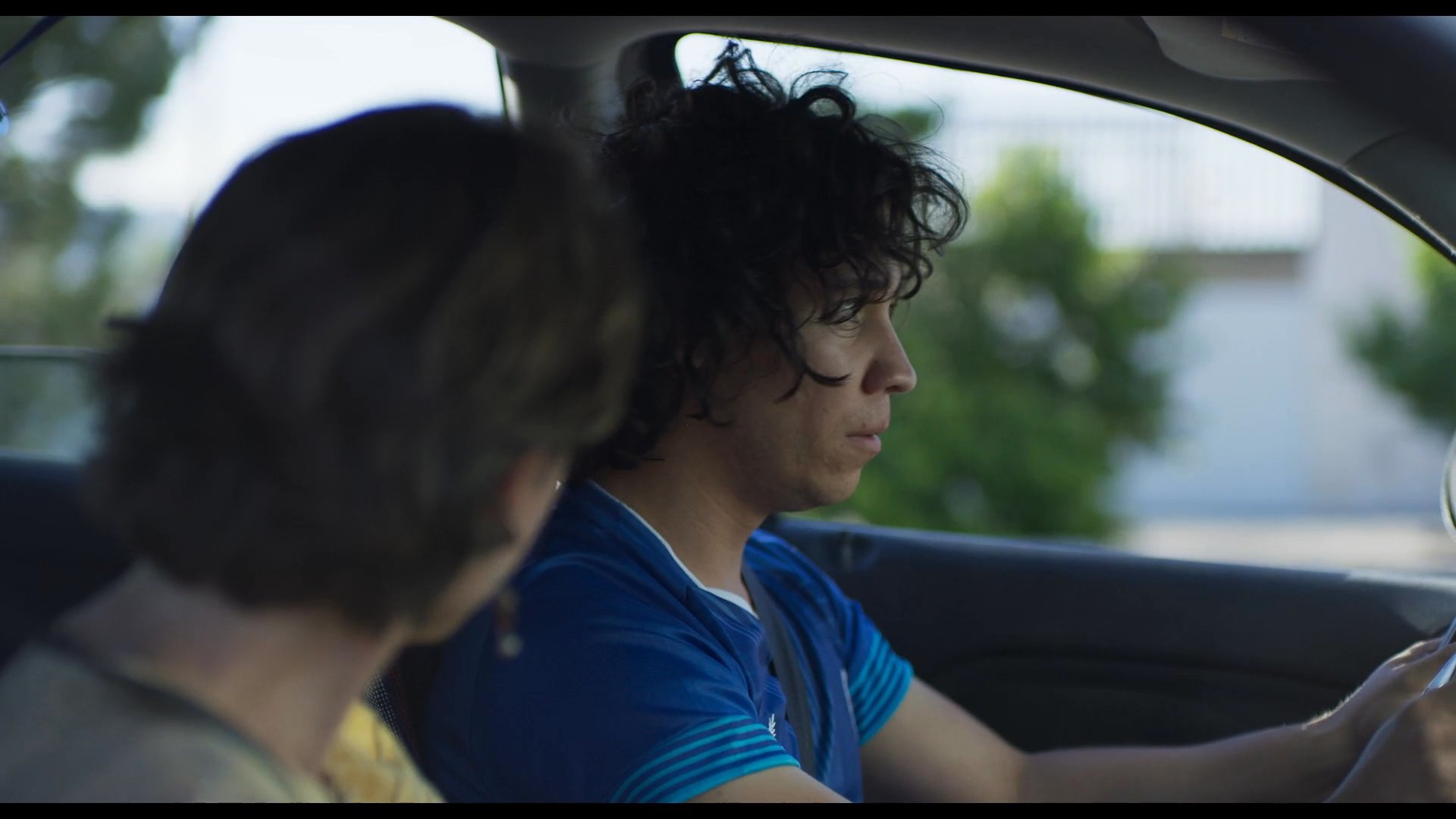 Two individuals engage in a focused, expressive conversation while seated in the front seats of a moving vehicle. Their facial expressions and body language suggest a dynamic exchange of thoughts during their commute.