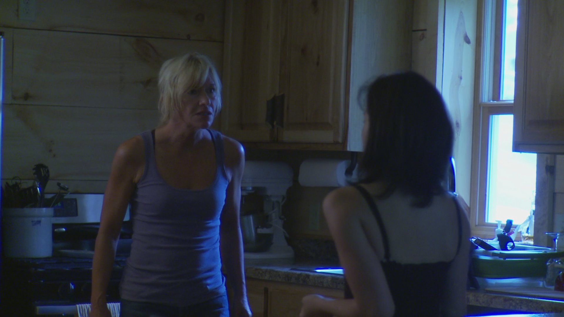 Two women engage in a tense, serious conversation inside a residential kitchen. The exchange involves expressive gestures and focused eye contact as they discuss a sensitive matter.
