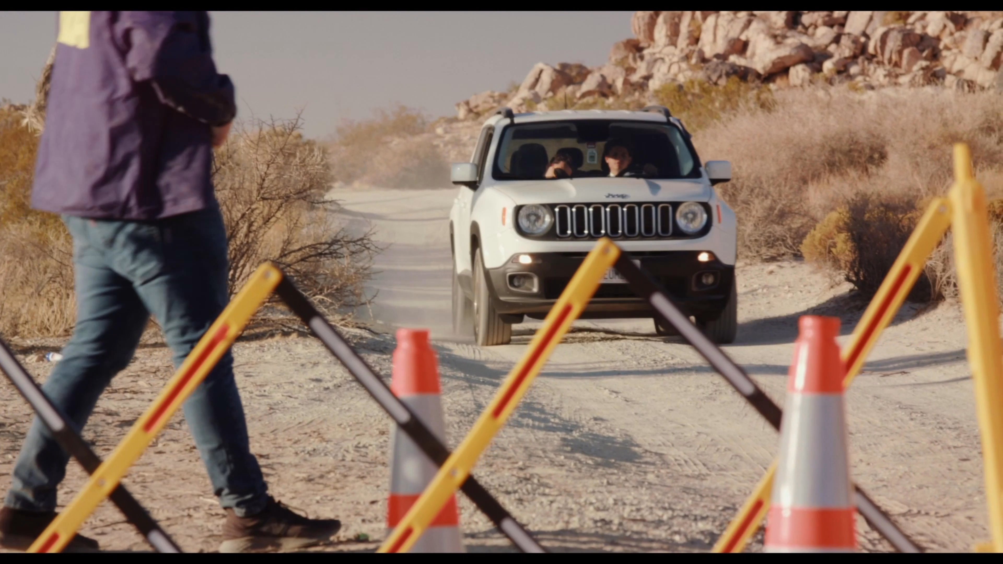 A DEA agent in a desert environment monitors an incoming white Jeep at a designated traffic checkpoint. The officer observes the vehicle as it approaches and pulls to a stop for inspection.