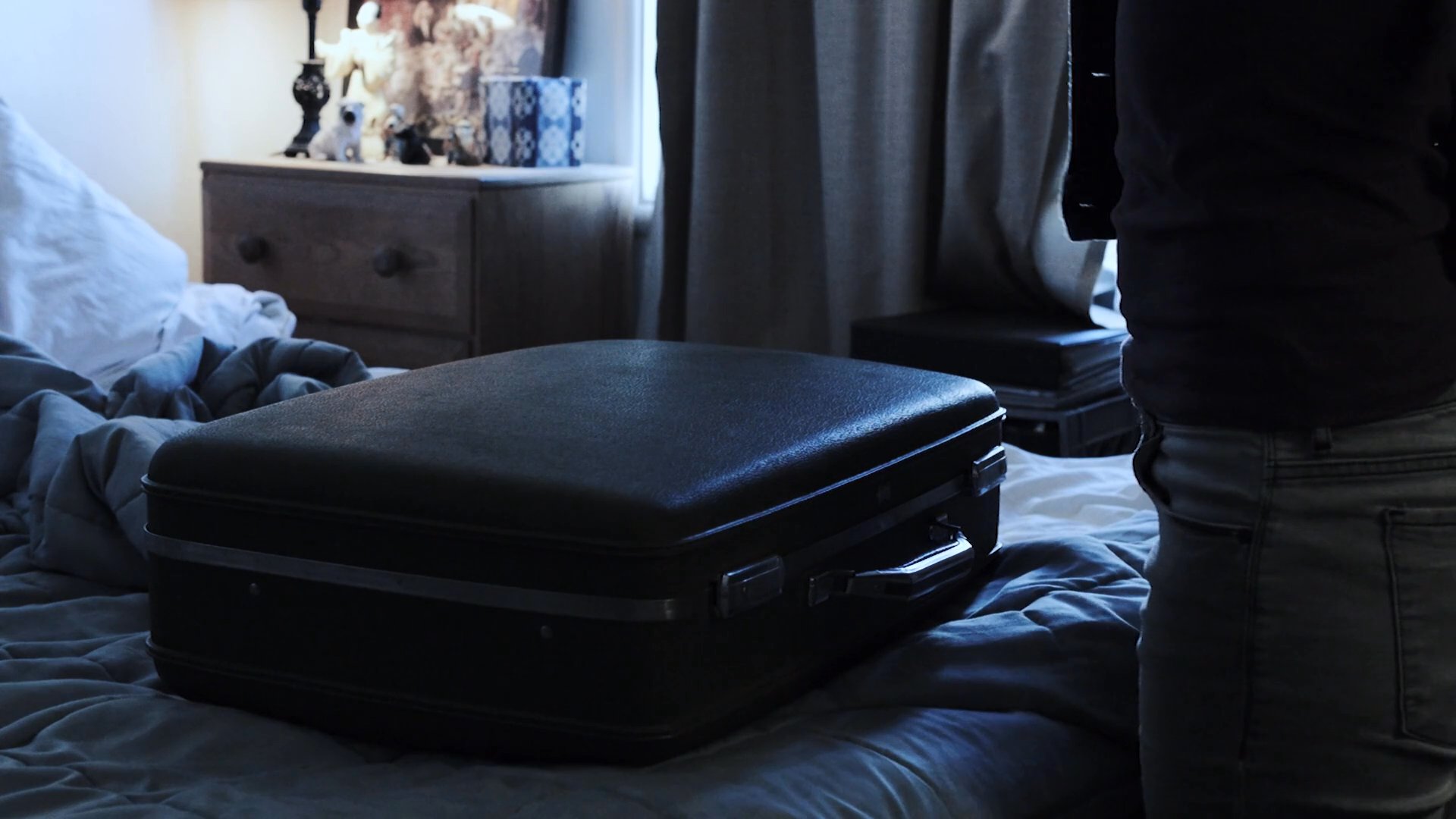 A person organizes a suitcase on a bed before counting a stack of cash taken from a cigar box.