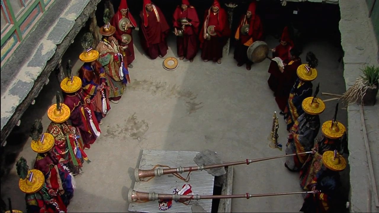 An elderly man describes traditions as monks perform a ritual within a remote Himalayan monastery courtyard. The scene captures the intersection of personal narrative and monastic life in a mountainous landscape.