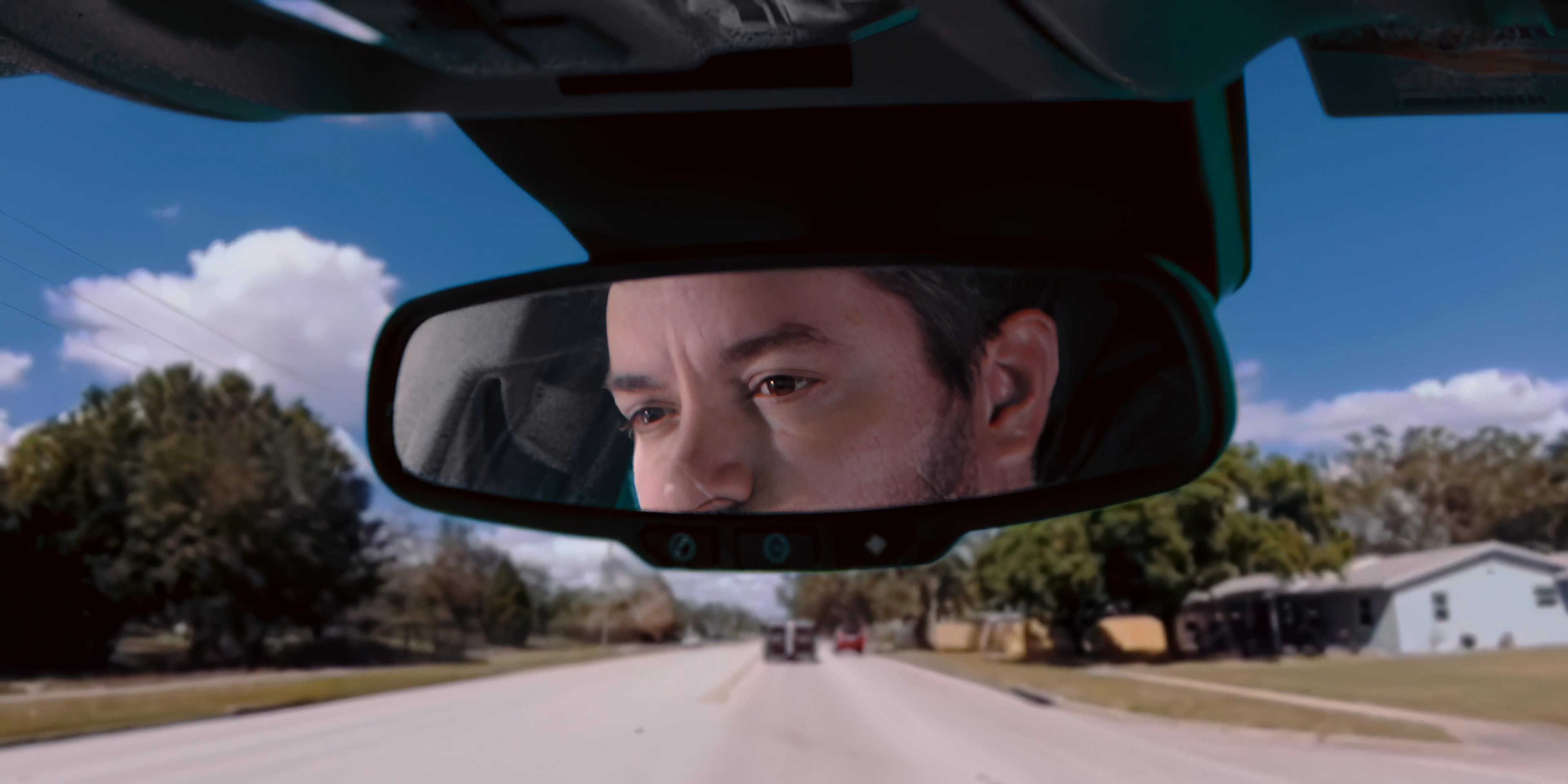 A man drives a vehicle while observing his sleeping passenger through the rearview mirror. The scene captures the quiet atmosphere of a daytime commute.