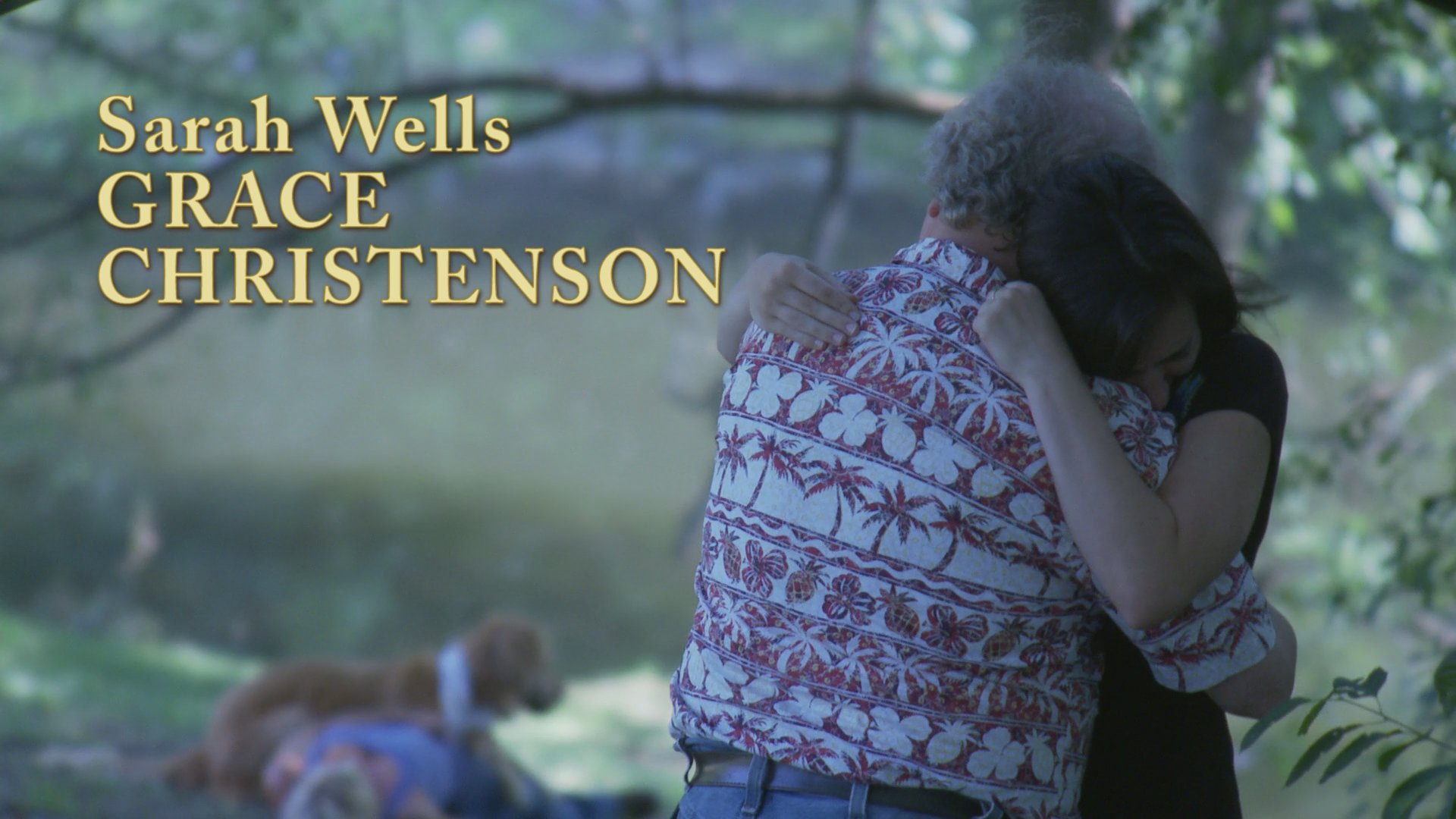 Two individuals share an intimate, sorrowful embrace by a lakeside while on-screen credits appear. A dog rests on the ground nearby in a somber, natural setting.