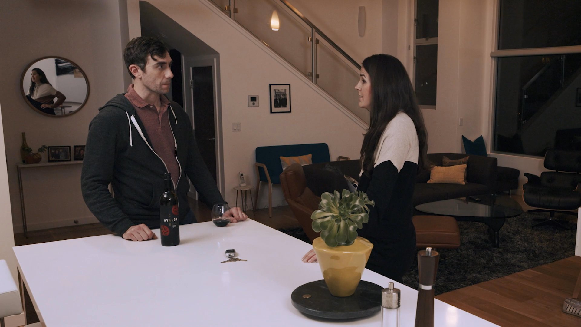 A man and woman engage in a tense, serious conversation while standing in a modern residential kitchen. The exchange focuses on emotional weight and interpersonal communication.