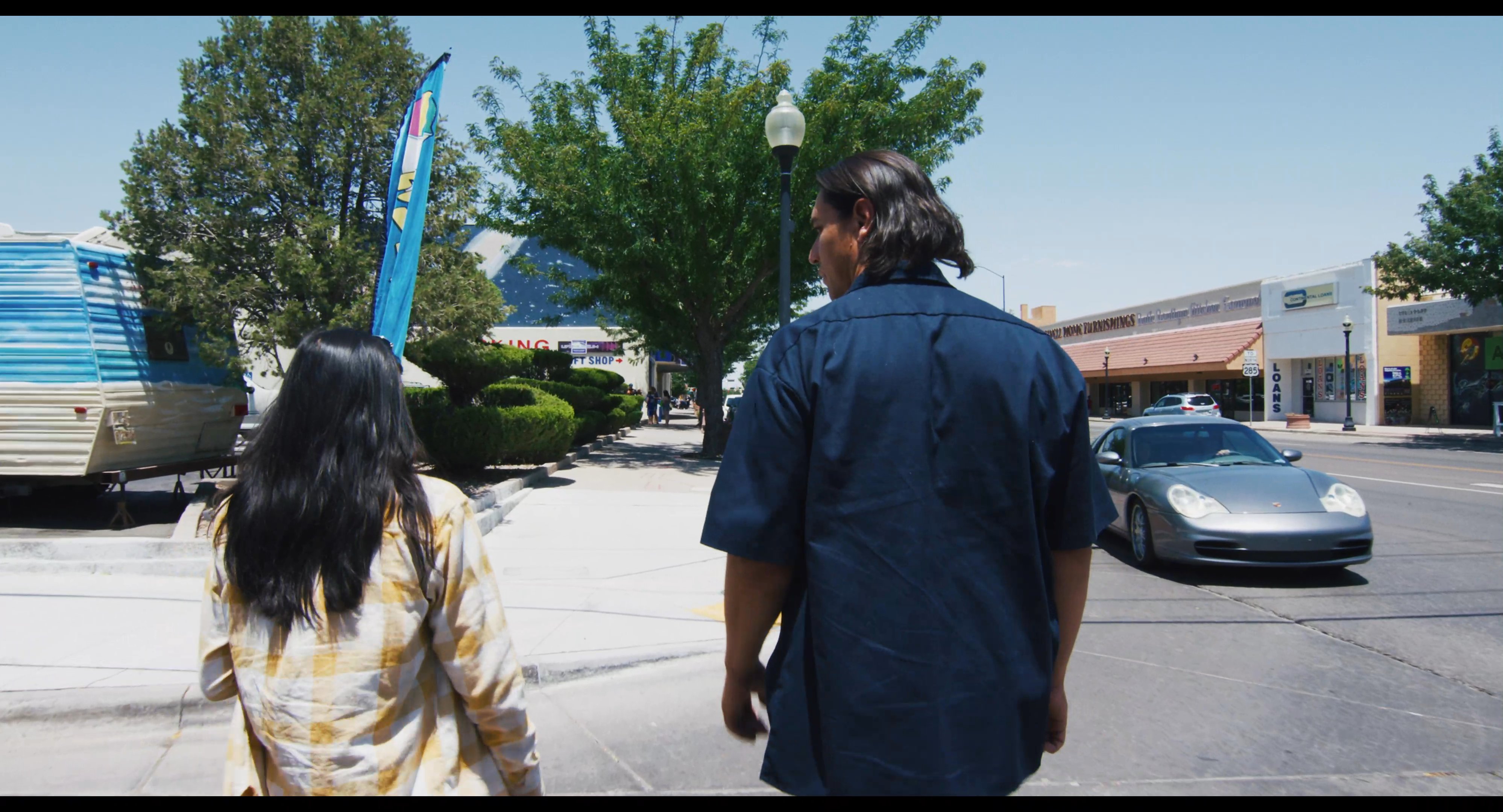 Two individuals walk together along an urban sidewalk near a local restaurant during the daytime. The scene captures their movement through a bustling street environment with active traffic.