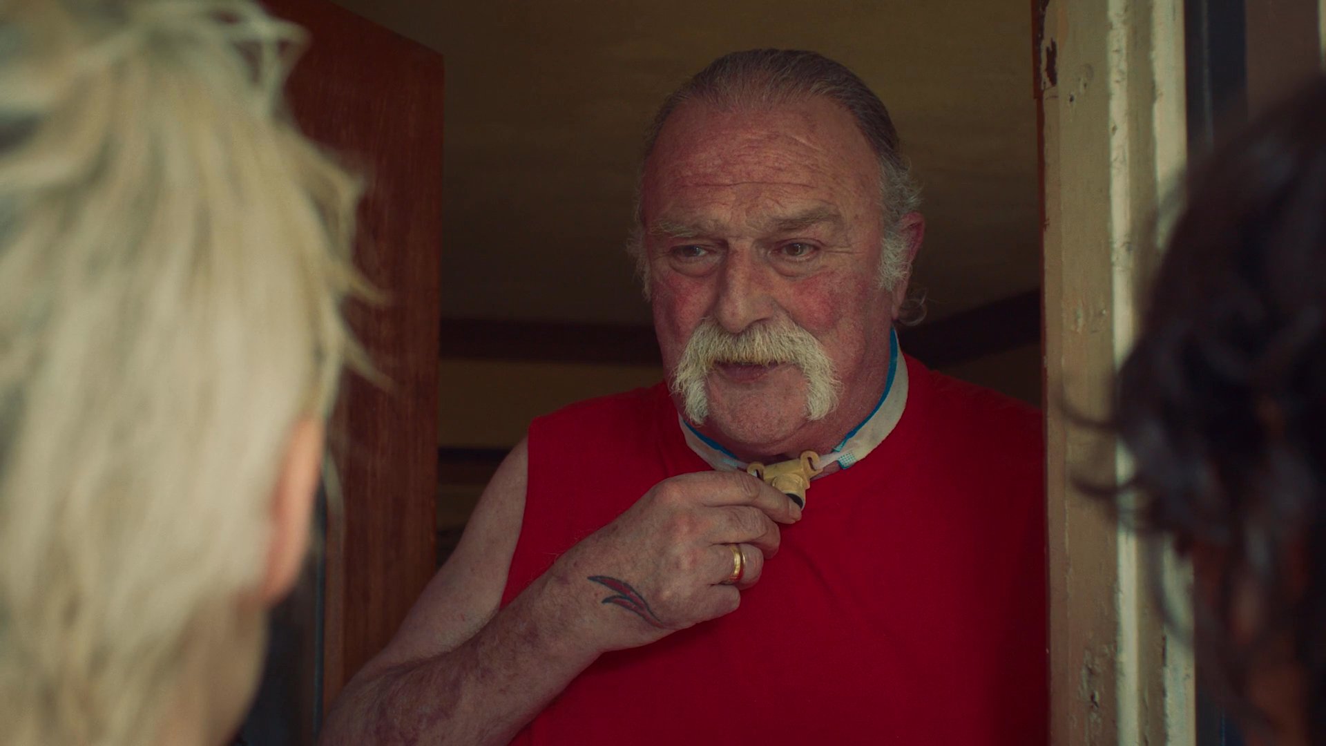 Two individuals approach a residence and engage in an interaction with a mustachioed man, who subsequently offers them a beverage inside his home.