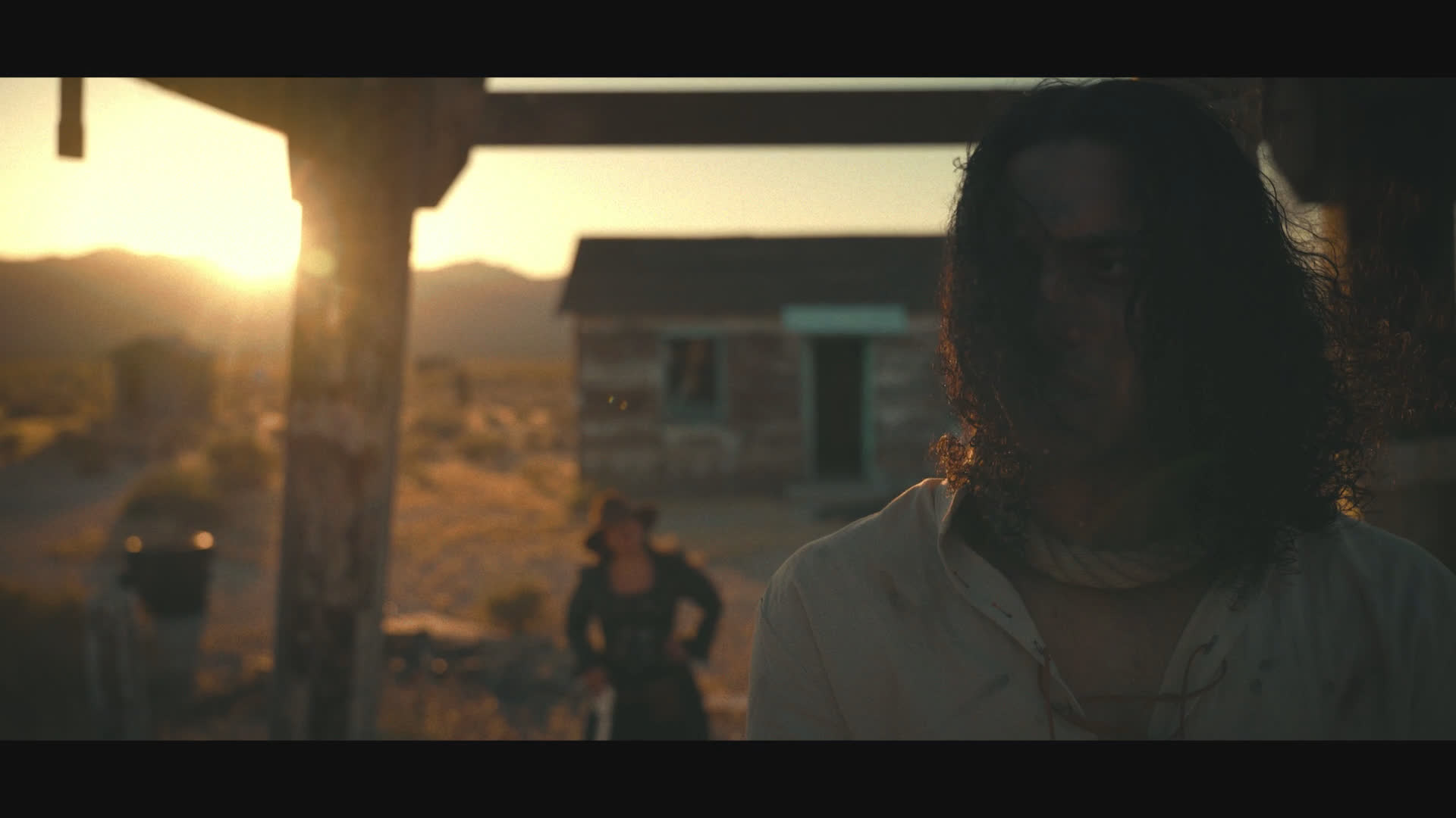 A long-haired man sits in the shadow of a doorway as a figure in a wide-brimmed hat approaches across a desolate homestead at sunset. They exchange tense questions about whether he needs help or is trying to kill himself.